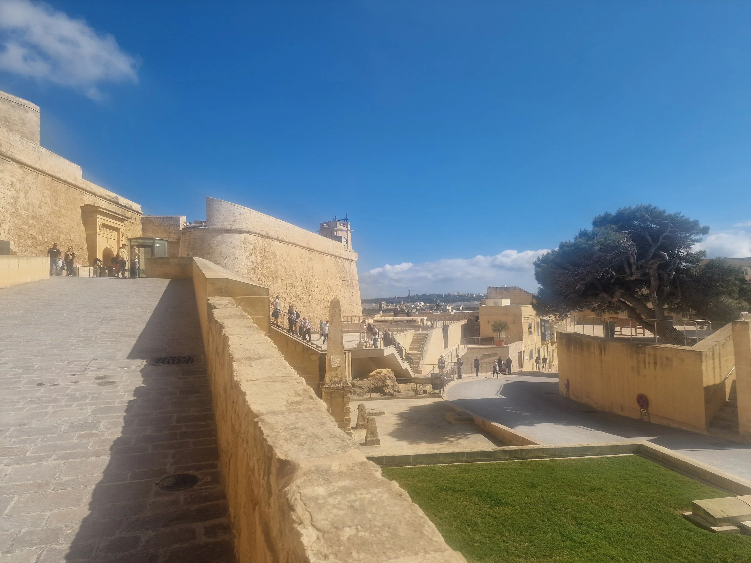 A historic stone fortress with people walking up and down the steps, a large green tree, and a bright blue sky with some clouds.