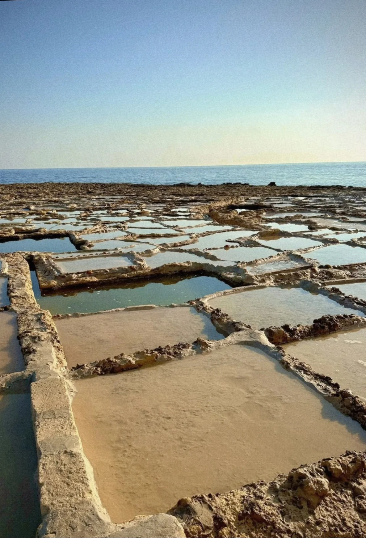 Gozo Malta , Salt flats or salt evaporation ponds near the ocean, with many rectangular shallow pools separated by salt-encrusted edges, under a clear sky.