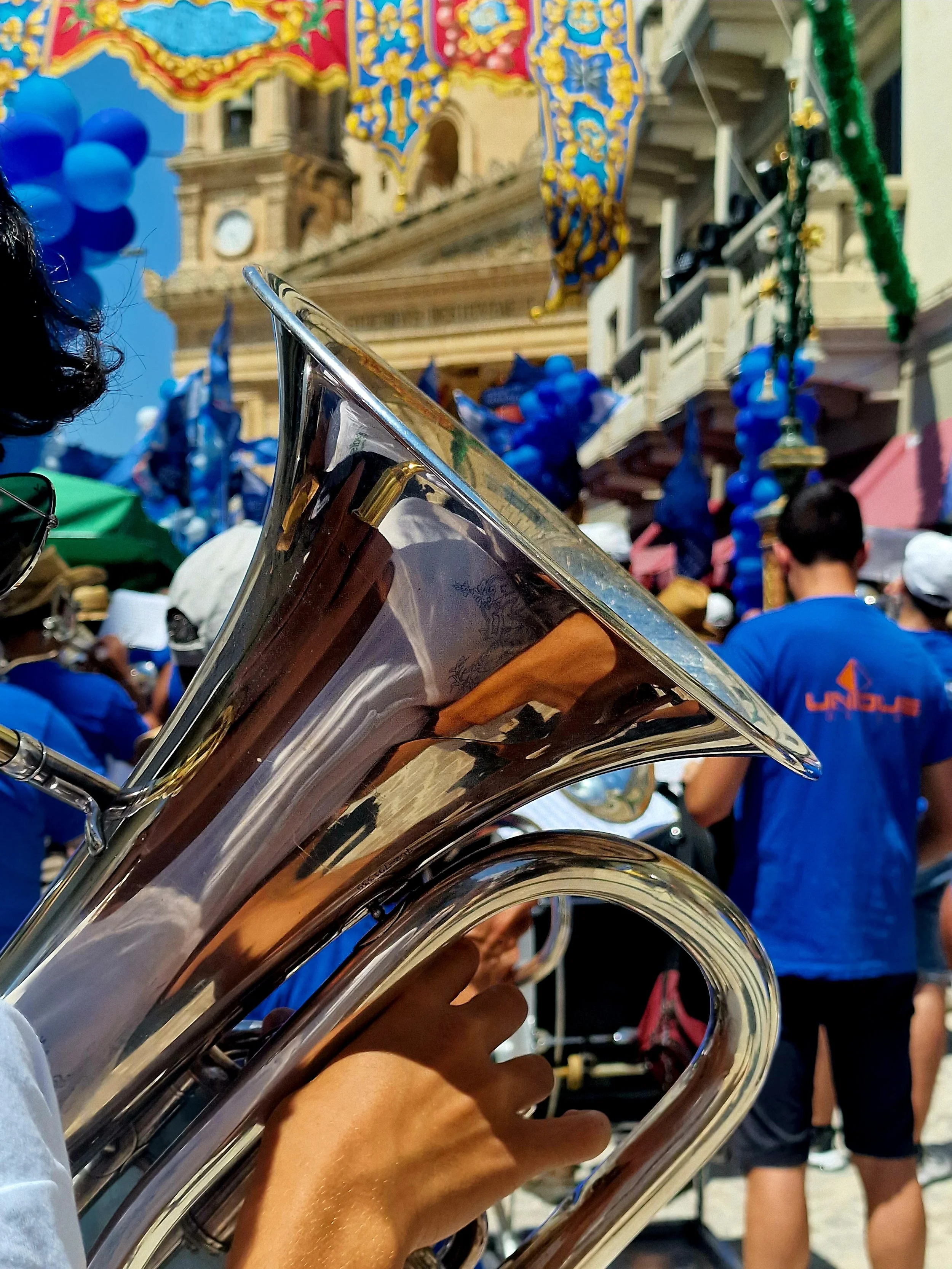 Close-up of a person's hand playing a silver trumpet, with a festive parade or celebration in the background featuring balloons, flags, a historical building, and people wearing blue shirts.