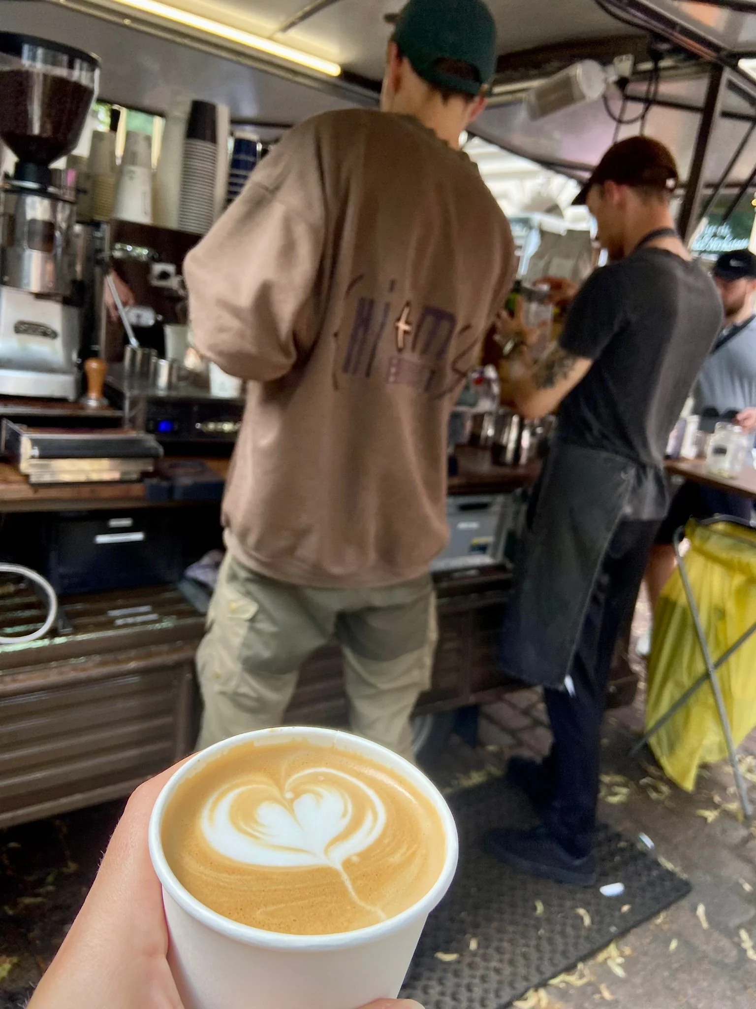 Hand hält eine Tasse mit Kaffee Latte, darauf ist ein Herz-Muster in Milchschaum. Im Hintergrund sind zwei Baristas in einer Kaffeebar zu sehen, die aus Kaffeemaschine und Utensilien arbeiten.