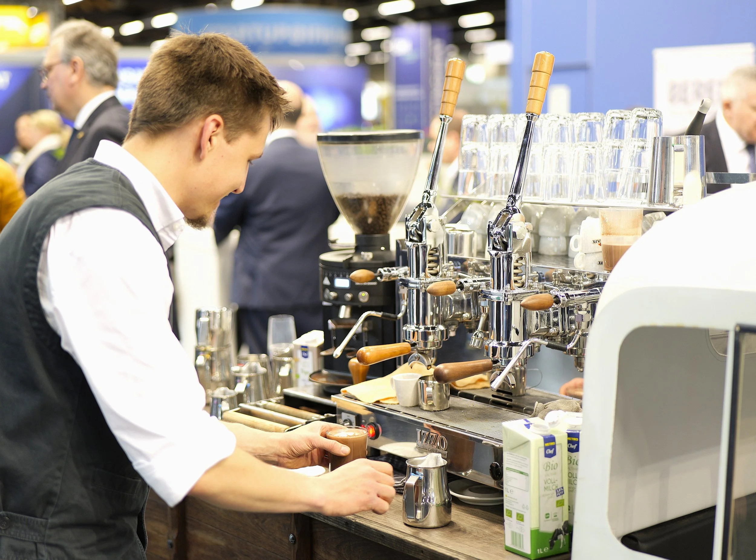 Ein Barista bereitet Kaffee in einer Espressomaschine zu in einem Kaffeeautomaten auf einer Messe oder einem Event, mit mehreren Gläsern und Verpackungen von Milch auf dem Tisch.