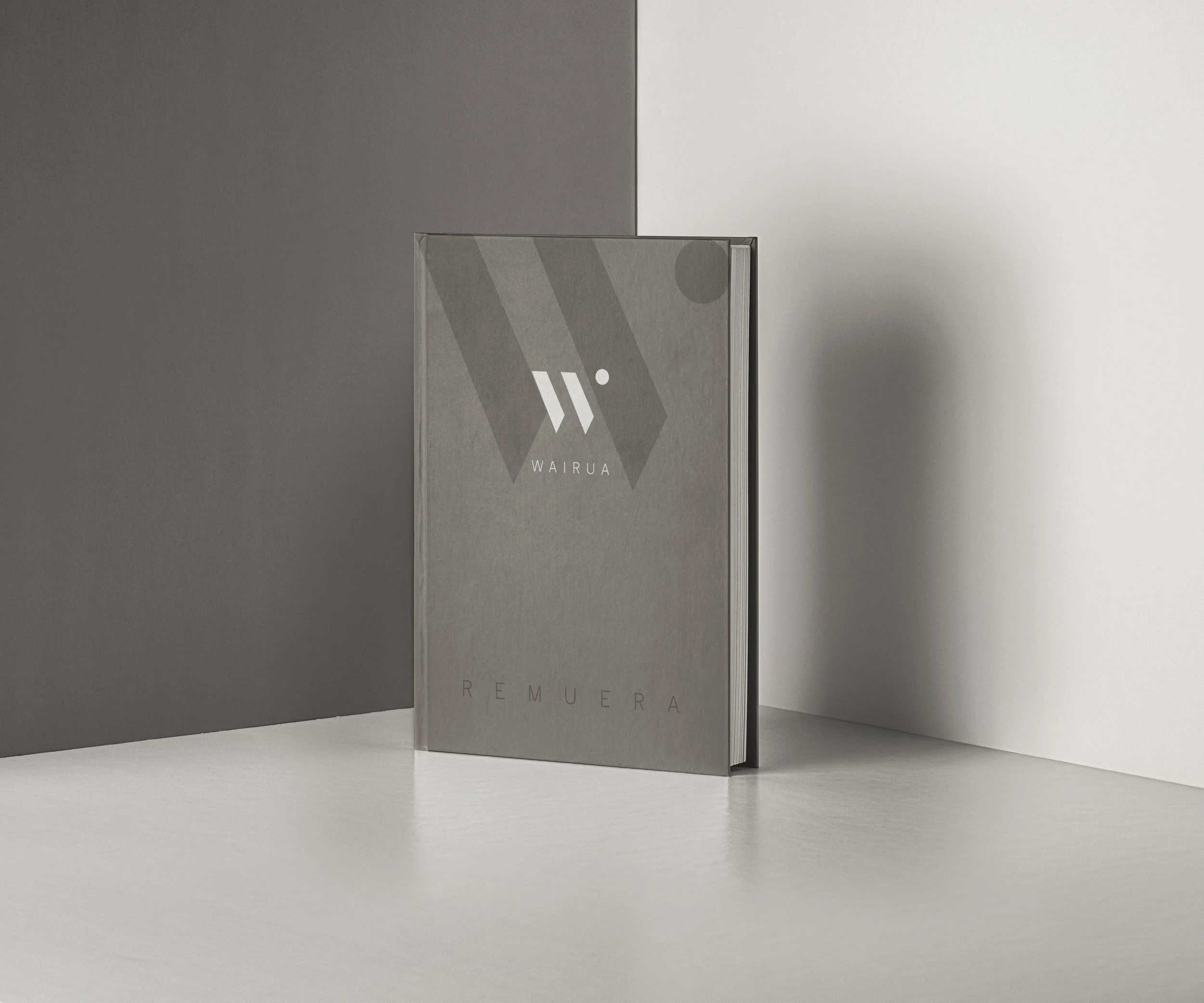 A gray book standing upright on a white surface against a gray and white background. The book cover features a minimalistic design with geometric shapes and the word 'REMUE' partially visible, and the word 'WAILUNA' at the bottom.