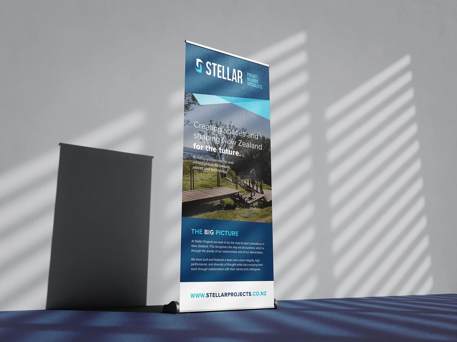 Standing banner with blue and white design promoting Stellar Projects, featuring a landscape photo of a person on a wooden path and information about their project focus. Shadows cast by window blinds are visible on the white wall background.