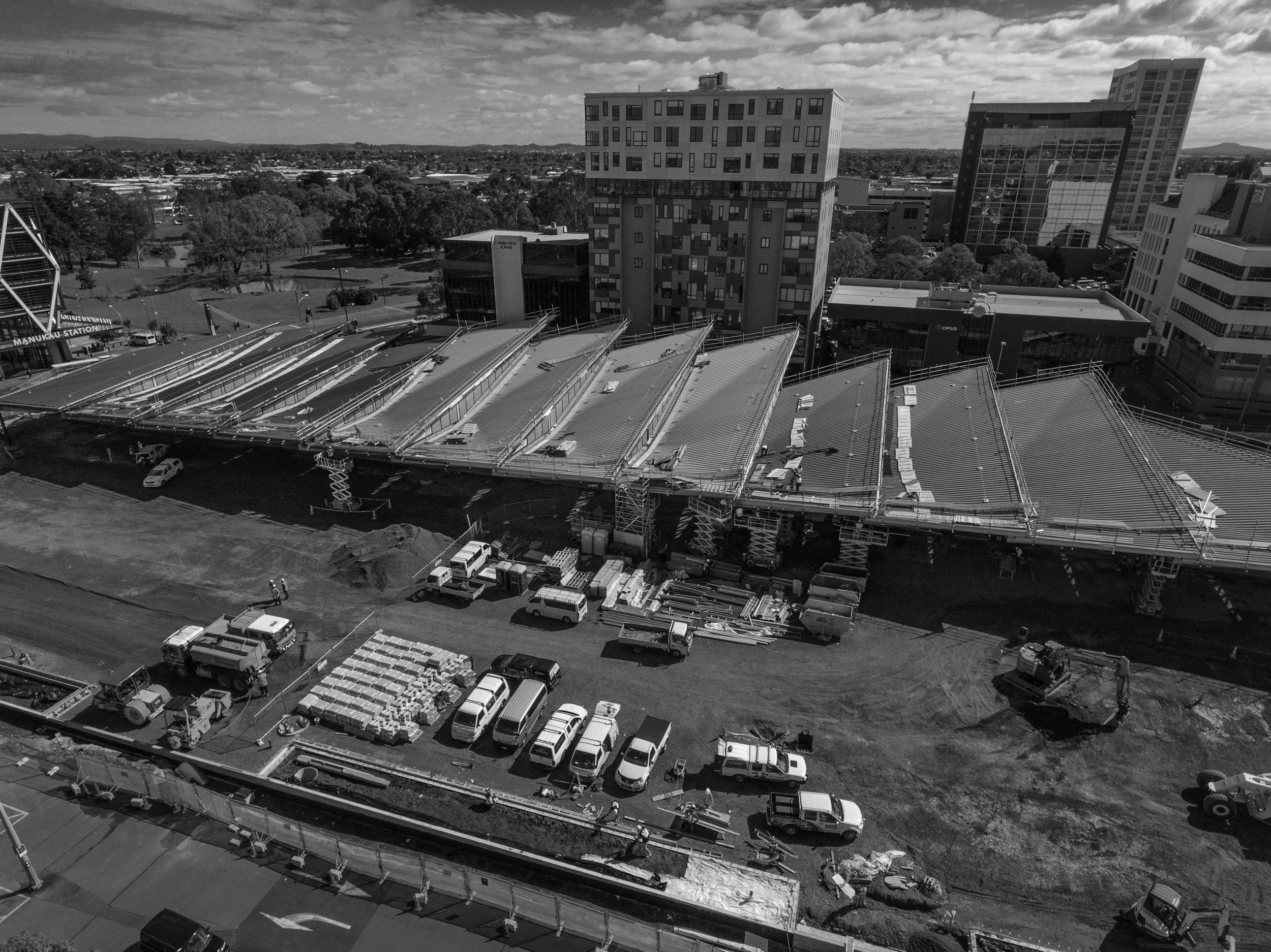 Construction site in an urban area with cranes, construction vehicles, and partially built structures, surrounded by high-rise buildings and a park.