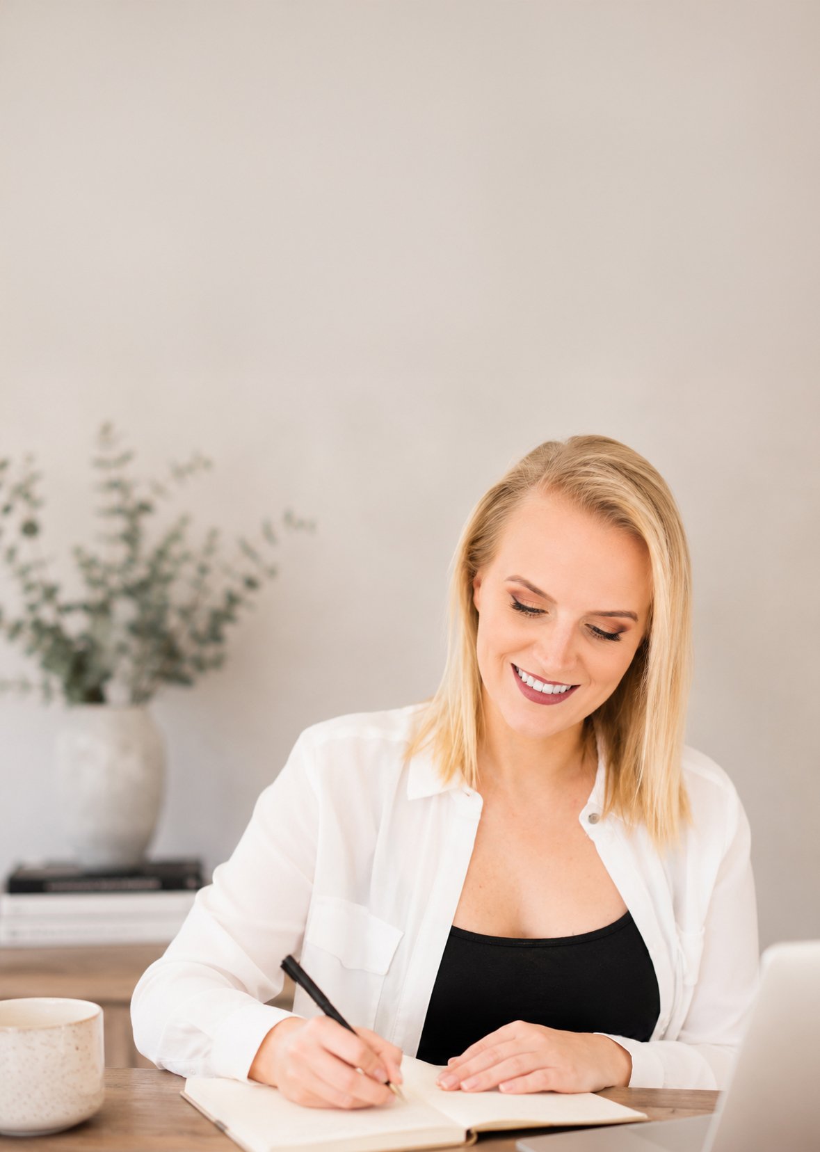 A woman with blonde hair smiling and writing in a notebook at a desk. In the background, there is a white vase with greenery on a table.