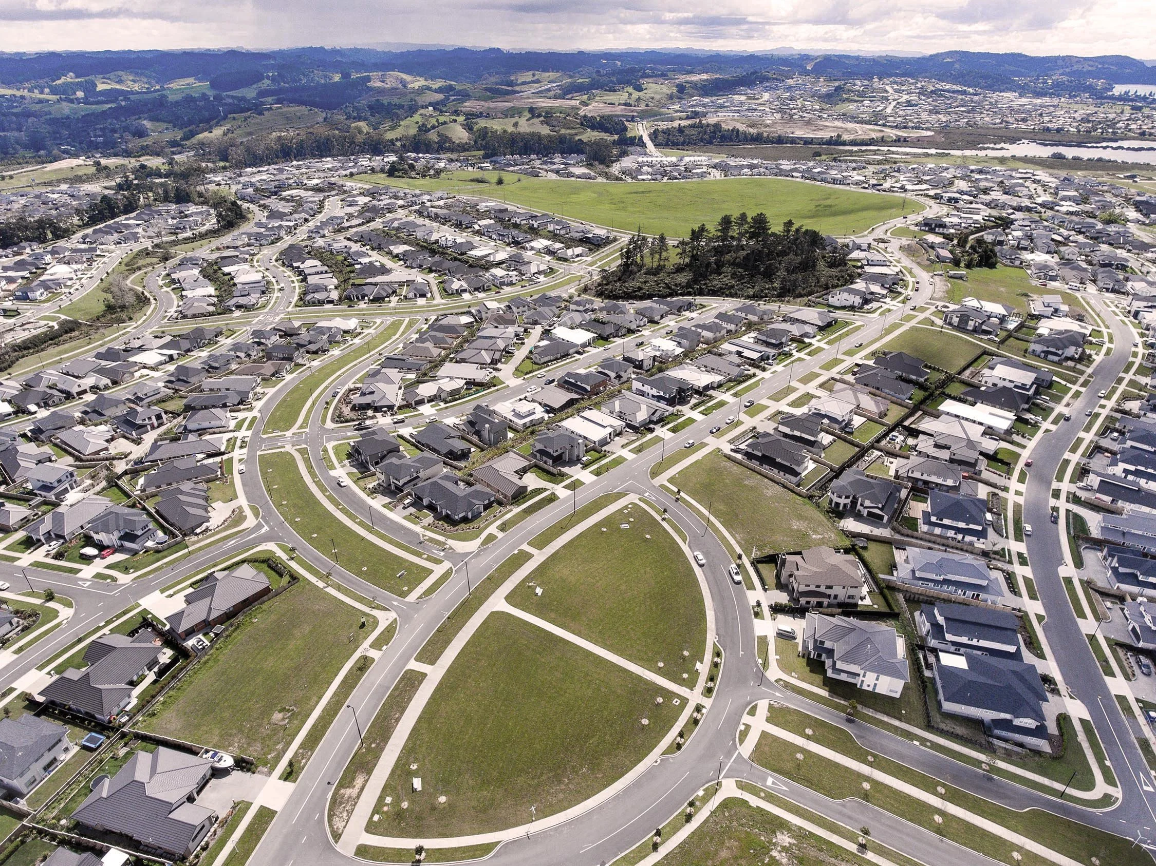 Aerial view of a residential neighborhood with houses, green lawns, and winding streets, surrounded by hills and a distant body of water.