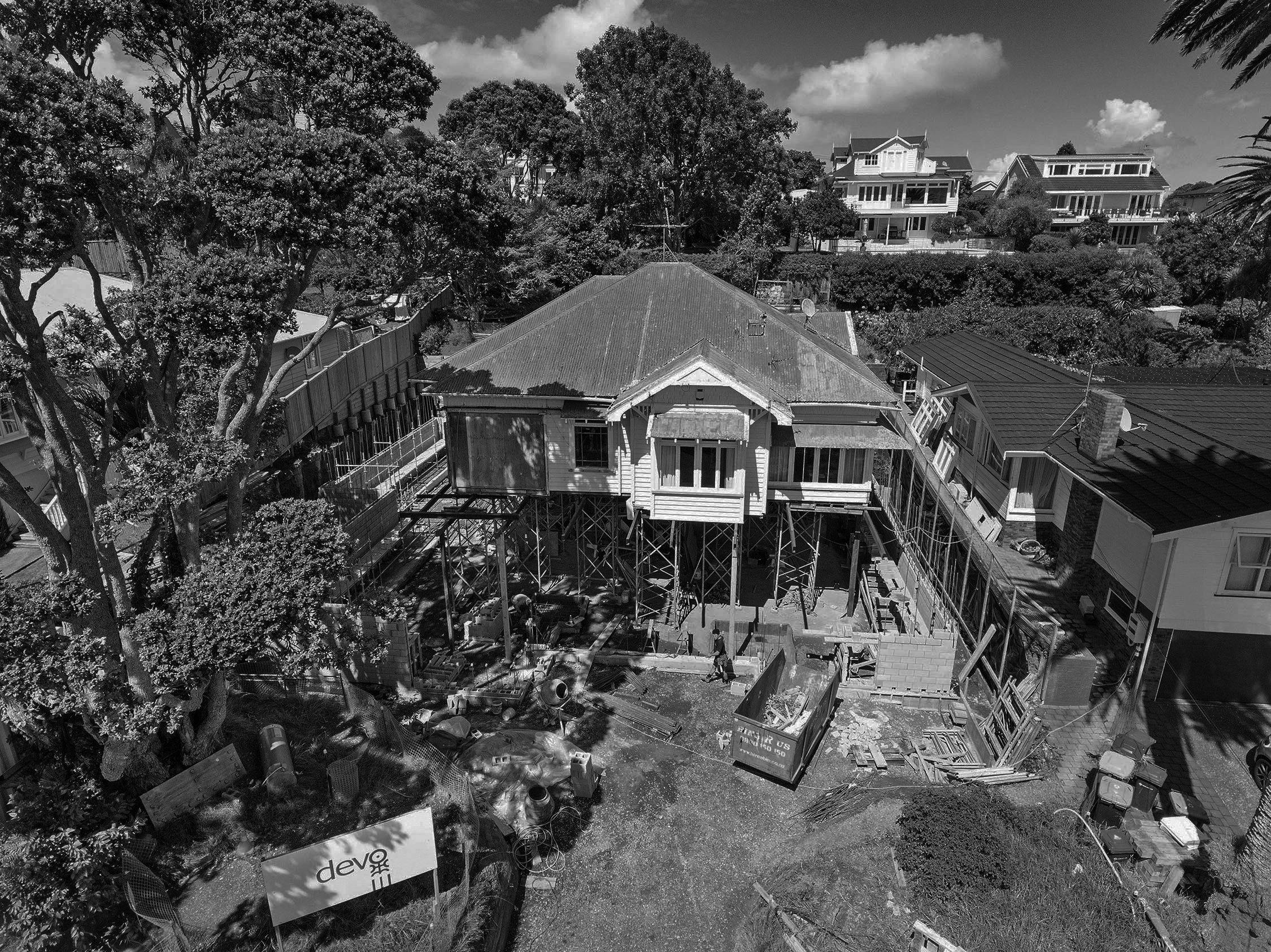 Black and white photo of a residential neighborhood with trees, houses, and a construction site in the foreground. There is scaffolding around one house, construction materials scattered around, and a sign that reads 'devo'.