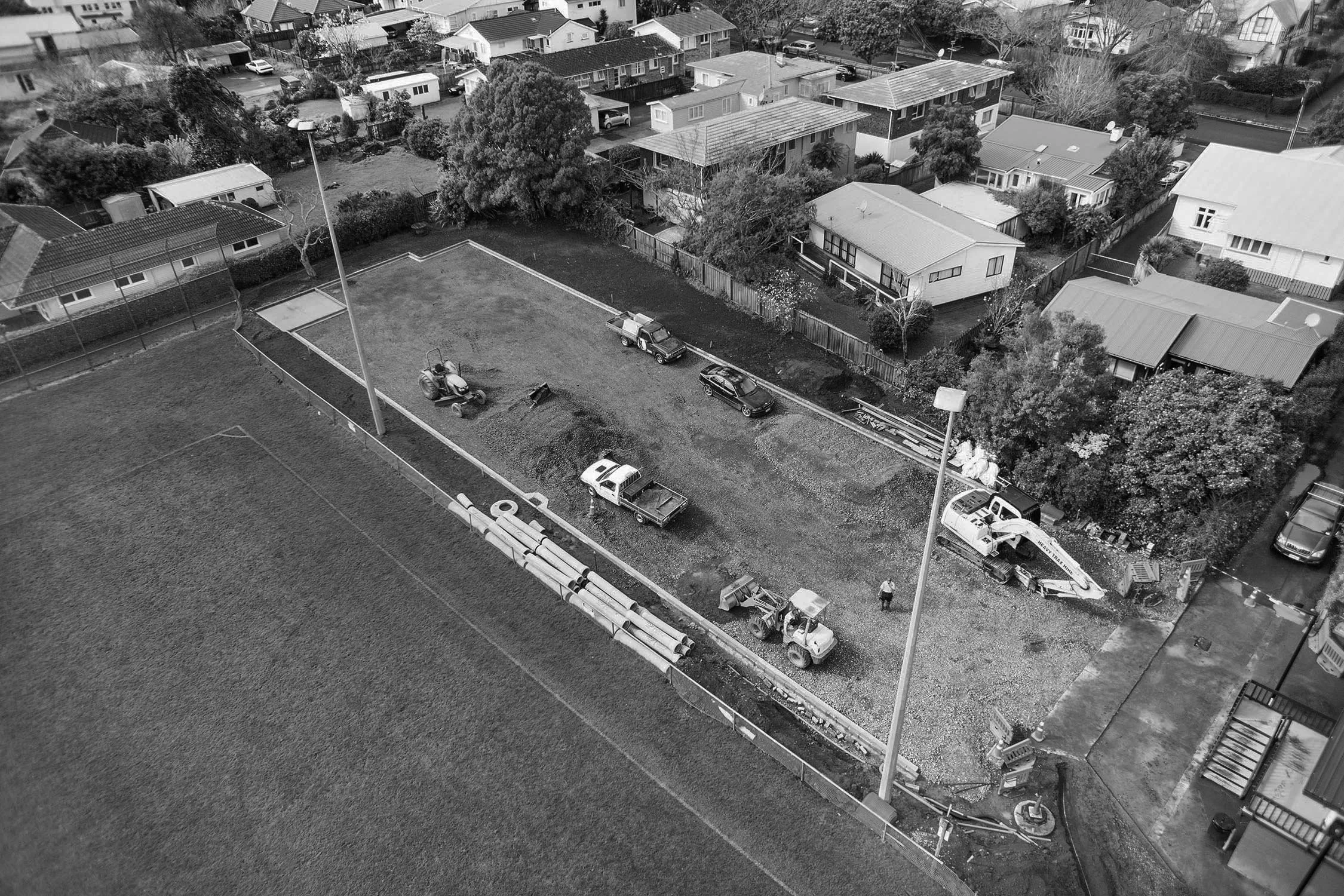A black and white aerial view of a residential neighborhood with houses and trees. In the foreground, there is a sports field under construction, with construction vehicles, equipment, and workers present.