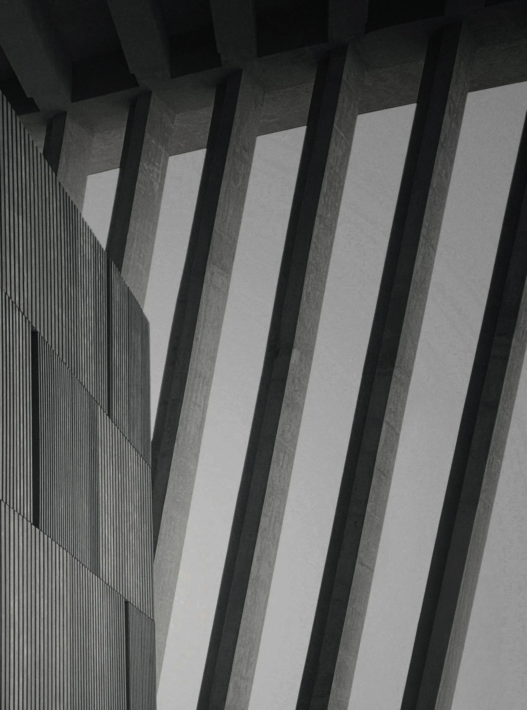 Close-up view of a modern building with large concrete and metal architectural elements, including vertical concrete beams and a ribbed metal facade, under a gray sky.