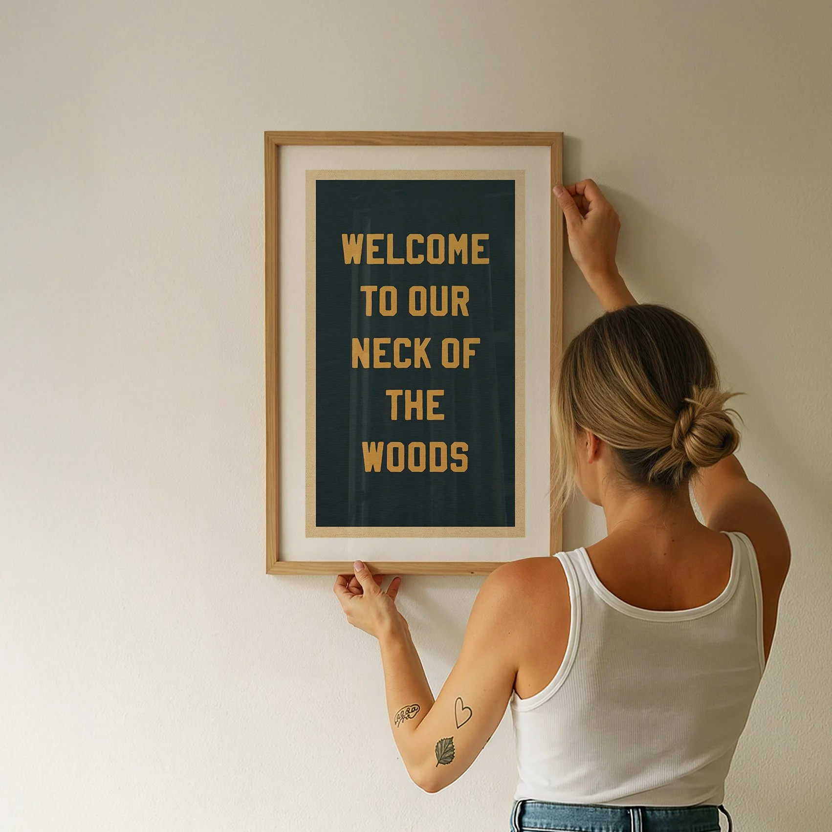 A woman holds a framed sign on a white wall that reads, "Welcome to our neck of the woods."