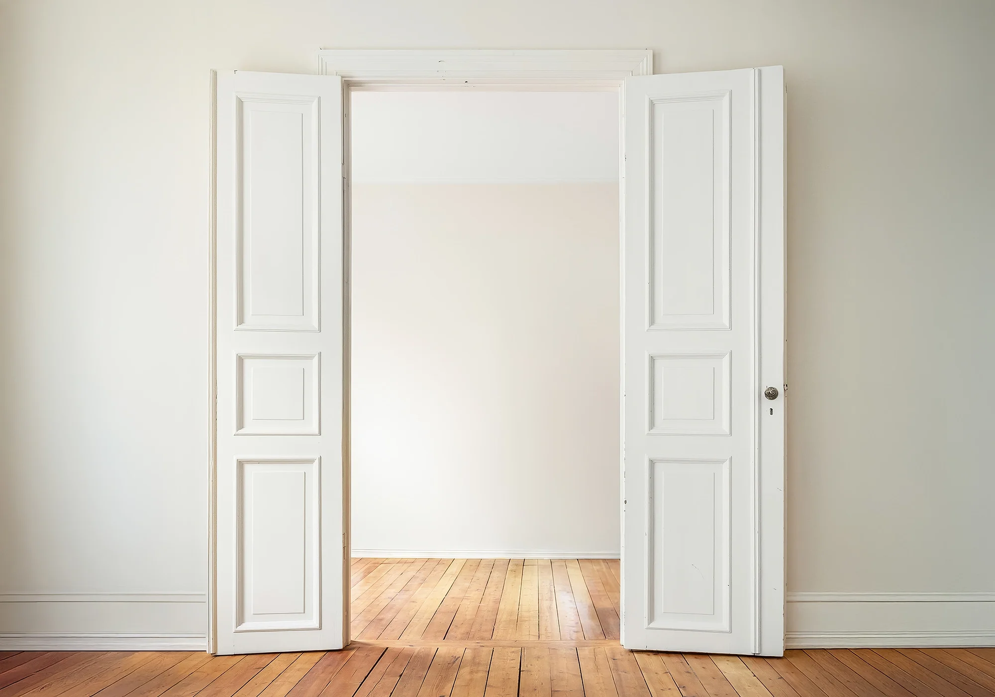 View of a white double door with open wooden shutters, leading to a bright room with wooden floor and beige walls.