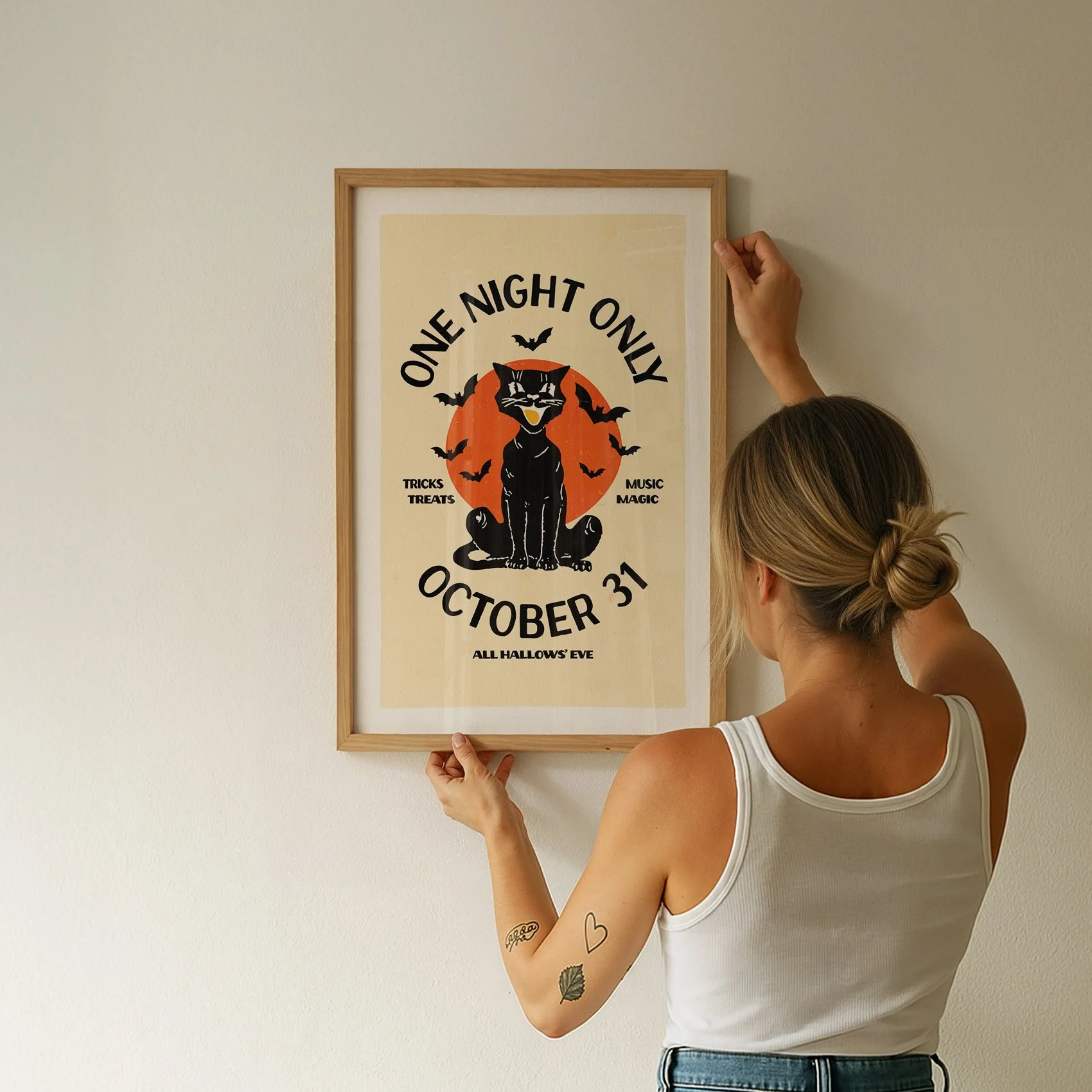 A woman with a tattoo on her right arm holds a framed Halloween-themed poster on a plain wall. The poster features a black cat, bats, and text about Halloween on October 31.