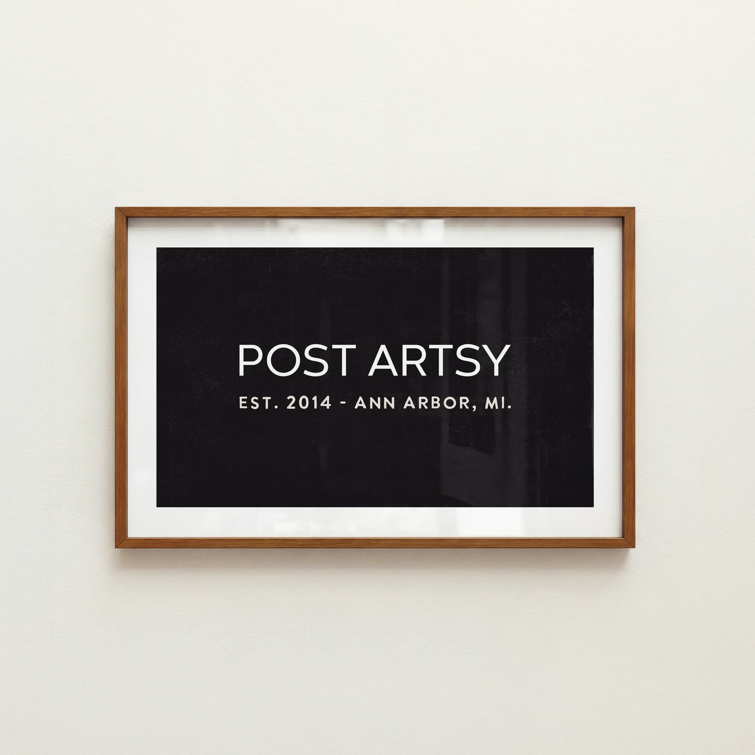 Framed black print with white text promoting brand PostArtsy from Ann Arbor, Michigan, on a white wall.