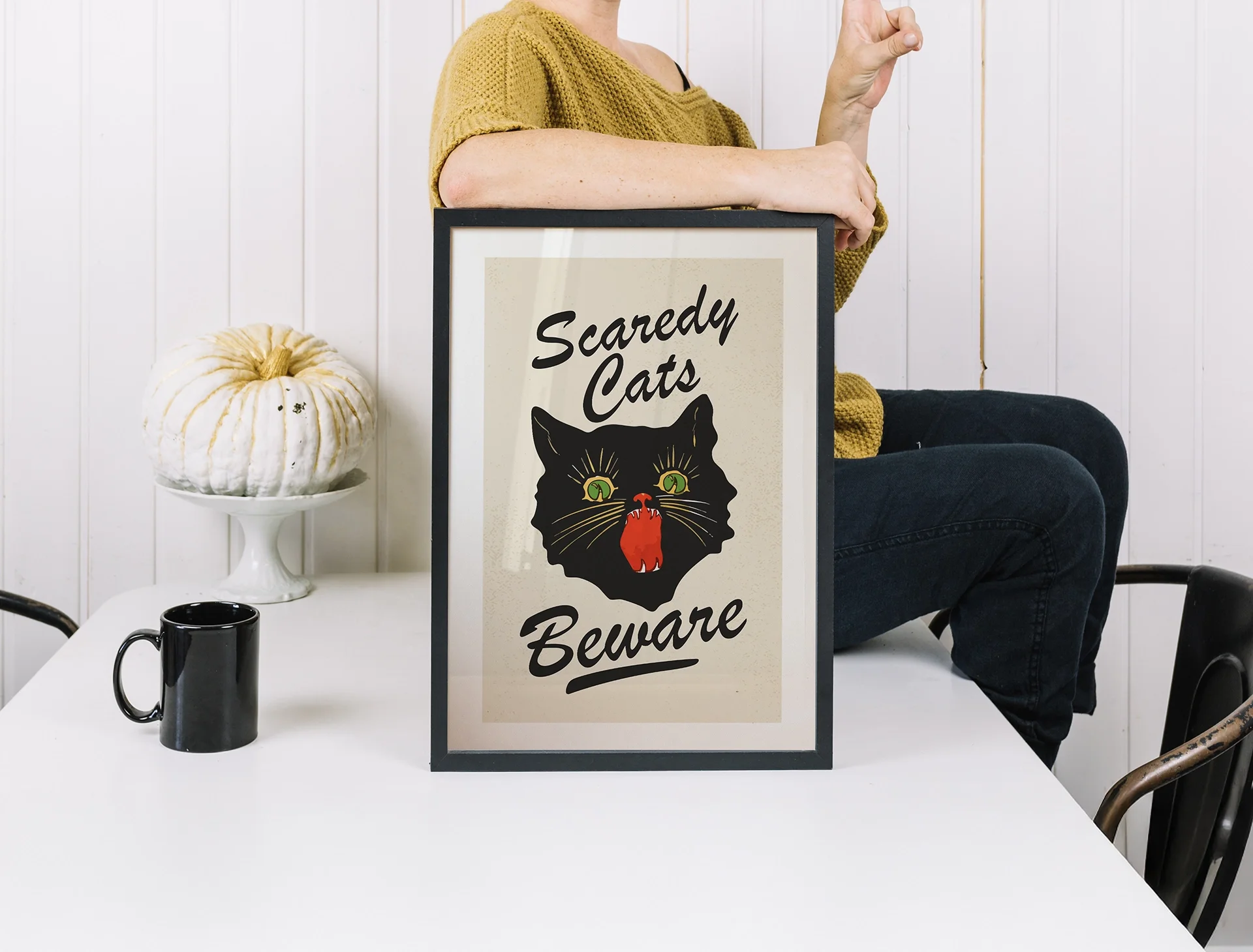 Person wearing a mustard-colored sweater sitting at a white table holding up a fist beside a sign that says "Scaredy Cats Beware" with a black cat illustration and a red mouth. A white pumpkin on a cake stand and a black mug are on the table, with a white paneled wall in the background.