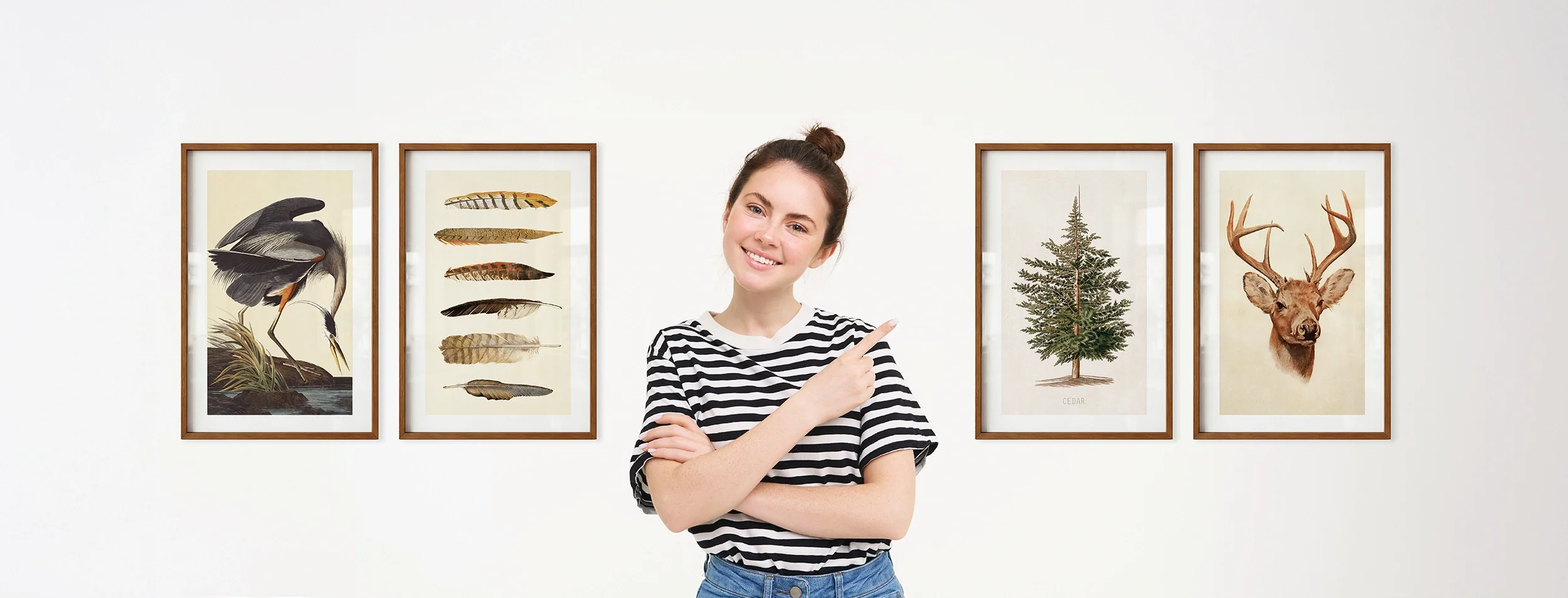 Young woman smiling and pointing at framed nature prints on a white wall, including a Blue Heron, bird feathers, a Cedar tree, and a deer with antlers.