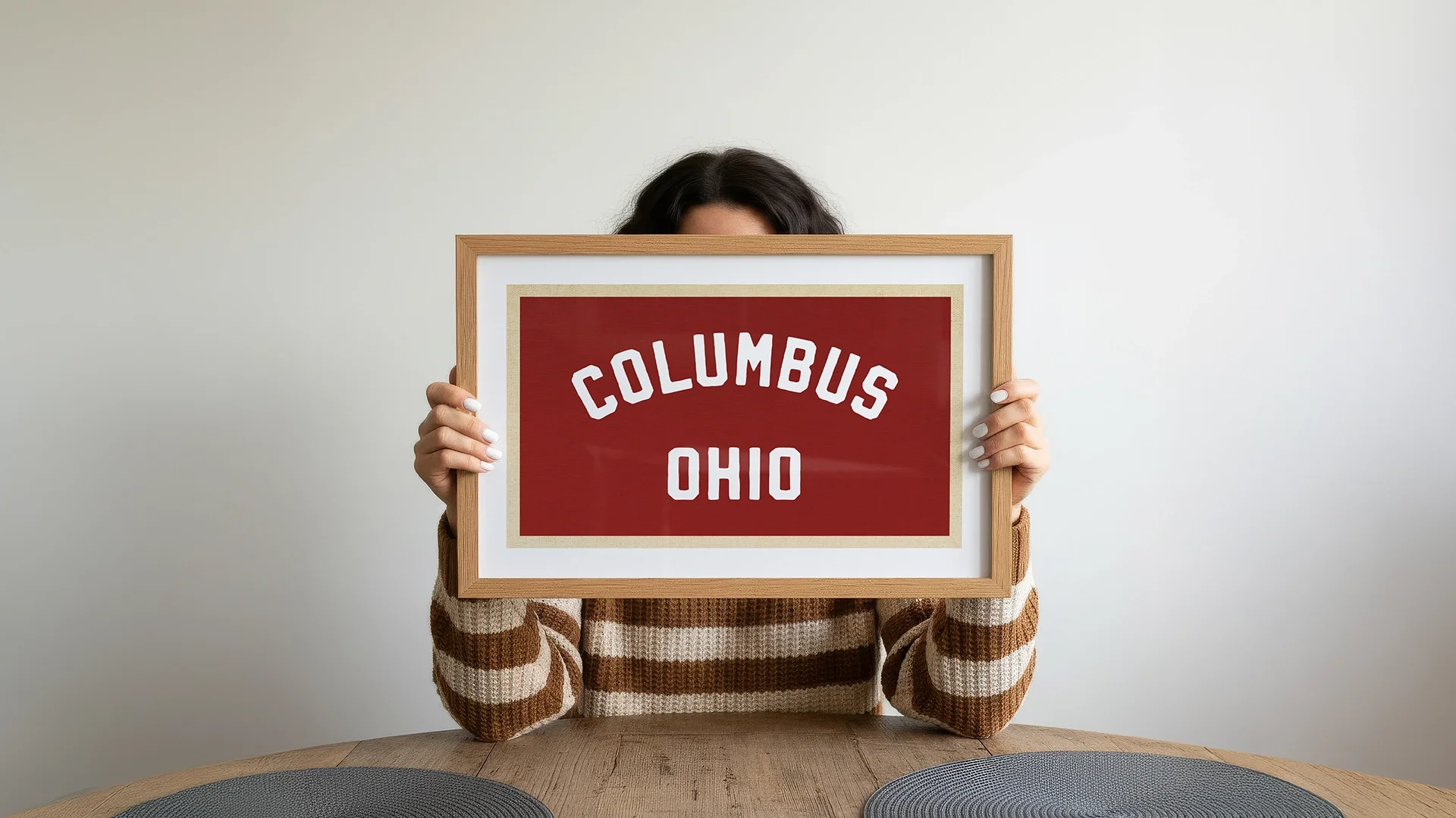 Person holding a framed sign that says 'Columbus Ohio' in front of their face, sitting at a table with two placemats.