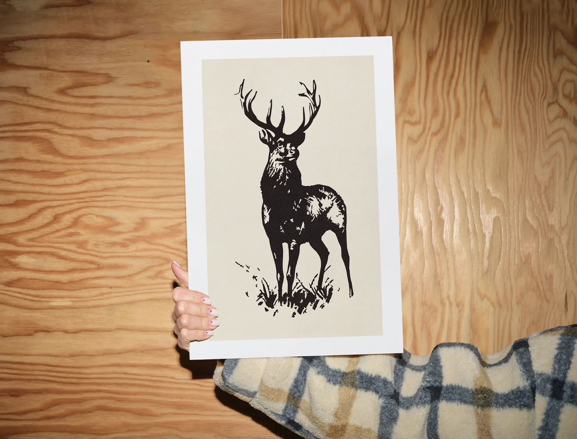 A person wearing a striped sweater holding a print of a black silhouette of a deer with large antlers on beige paper, against a wooden background.