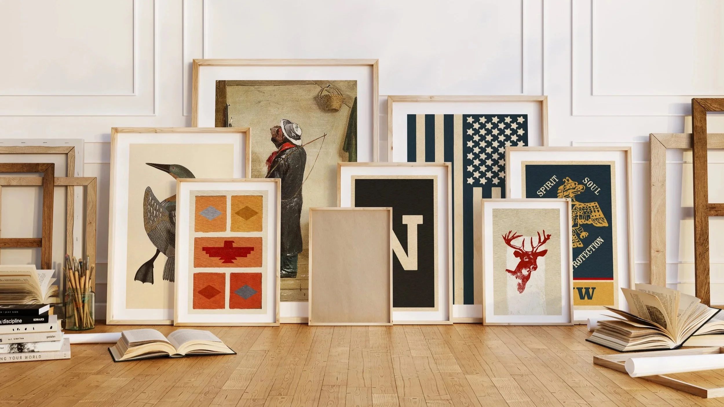 A display of various framed posters and artwork leaning against a white wall, with open books and rolled papers on the floor.