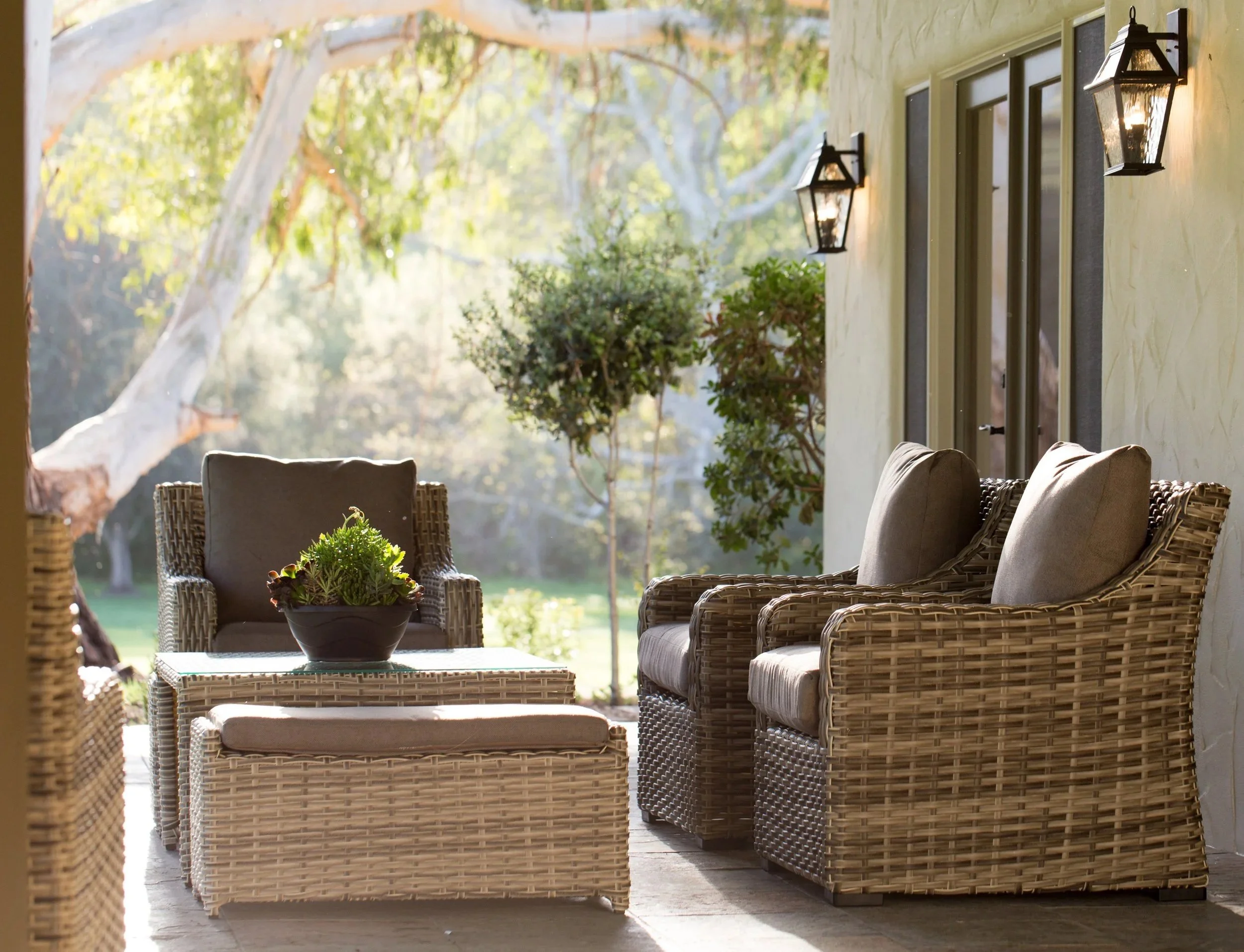 Outdoor patio area with wicker chairs and loveseats, potted plants, and large windows, overlooking a yard with trees and greenery.