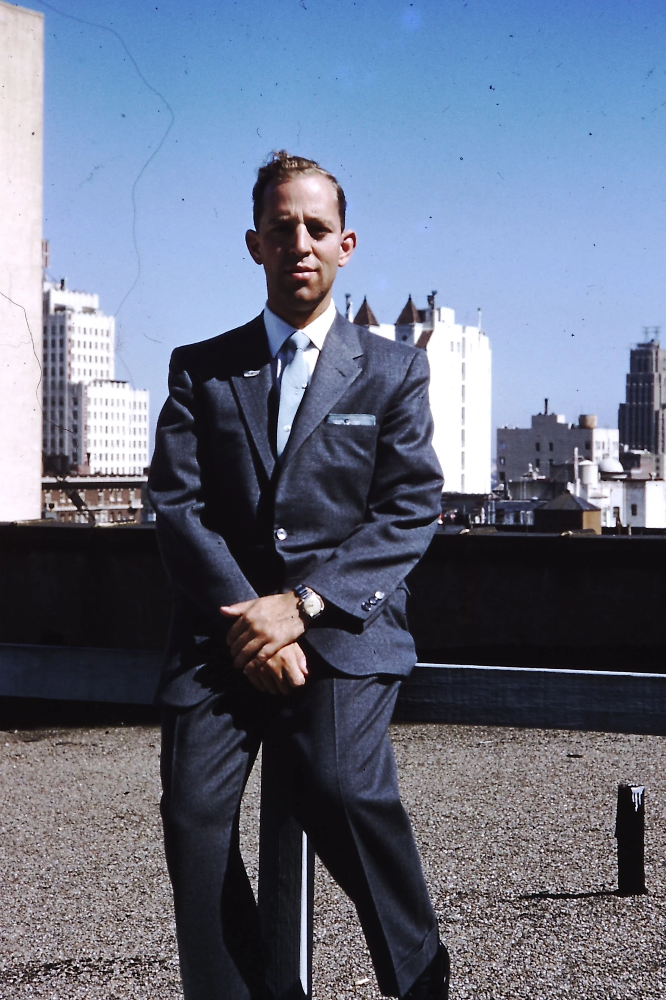 Spencer Maxwell in a gray suit standing outdoors on a rooftop with city skyscrapers in the background, wearing a wristwatch and a tie.