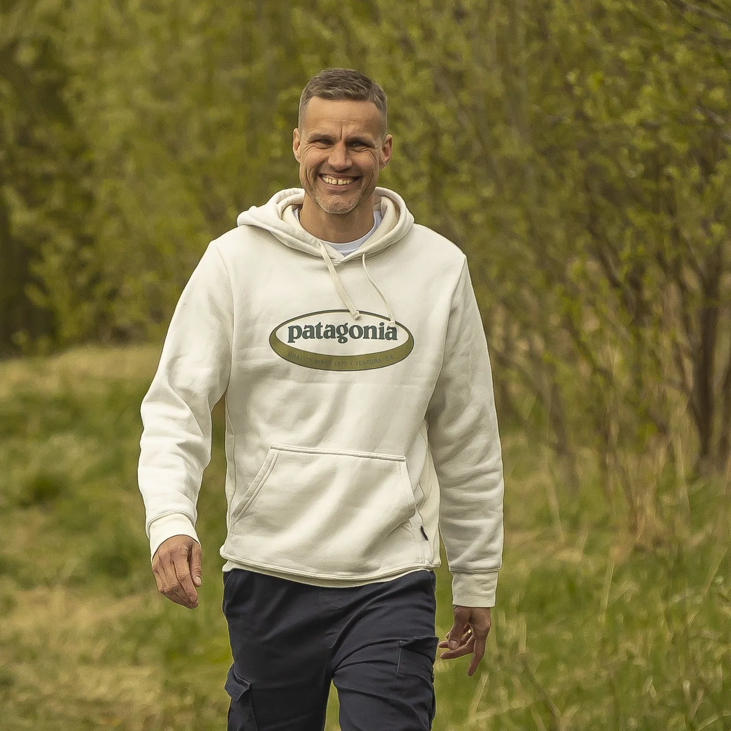 En mann som går utendørs, smilende, iført en hvit hoodie med Patagonia-logo, omgitt av grønne trær og gress.