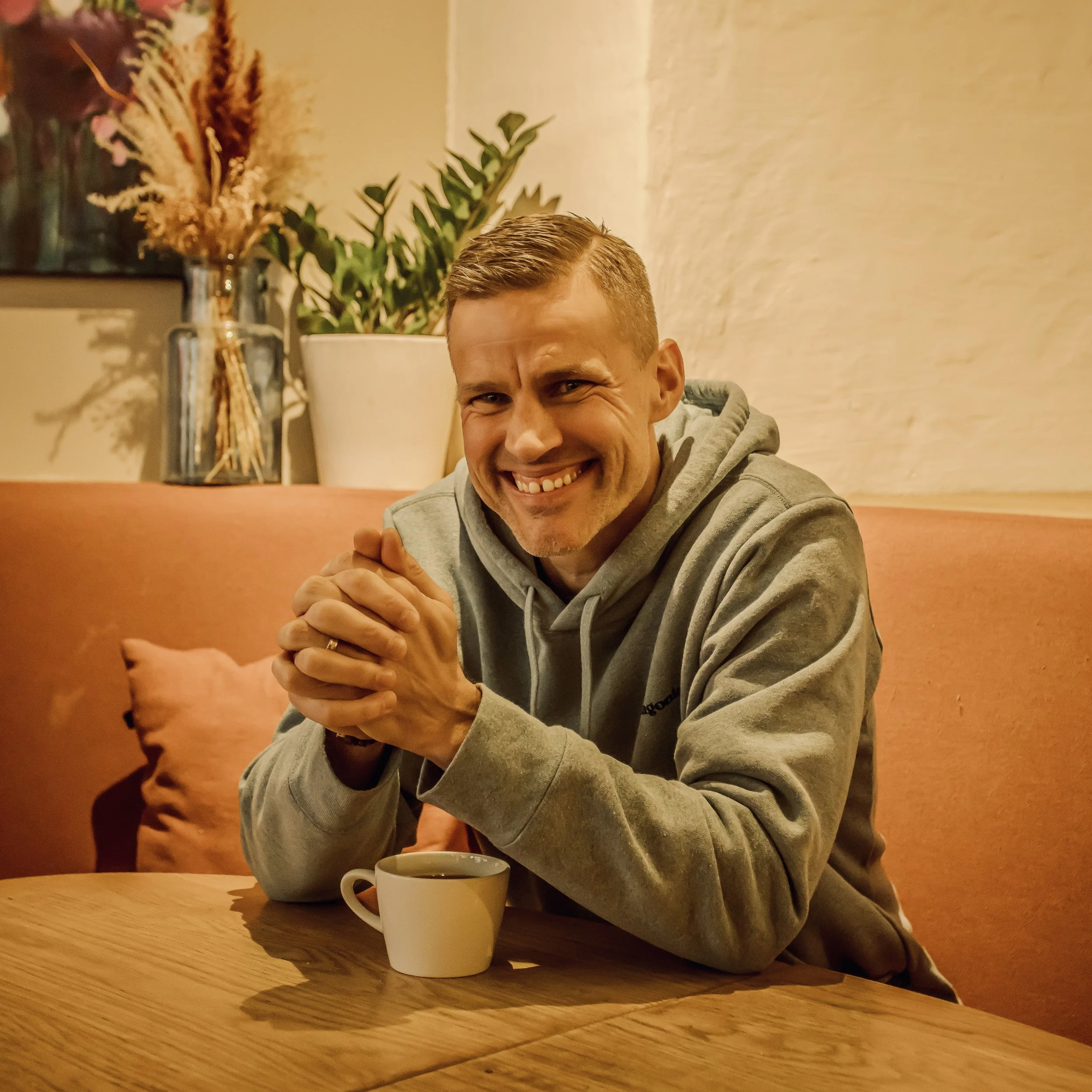 Et smilende man med kort hår og grågenser sitter ved et trebord med en hvit kopp foran seg, i en varm innredningsstil med en plante og en blomsterbukett i vaske i bakgrunnen.