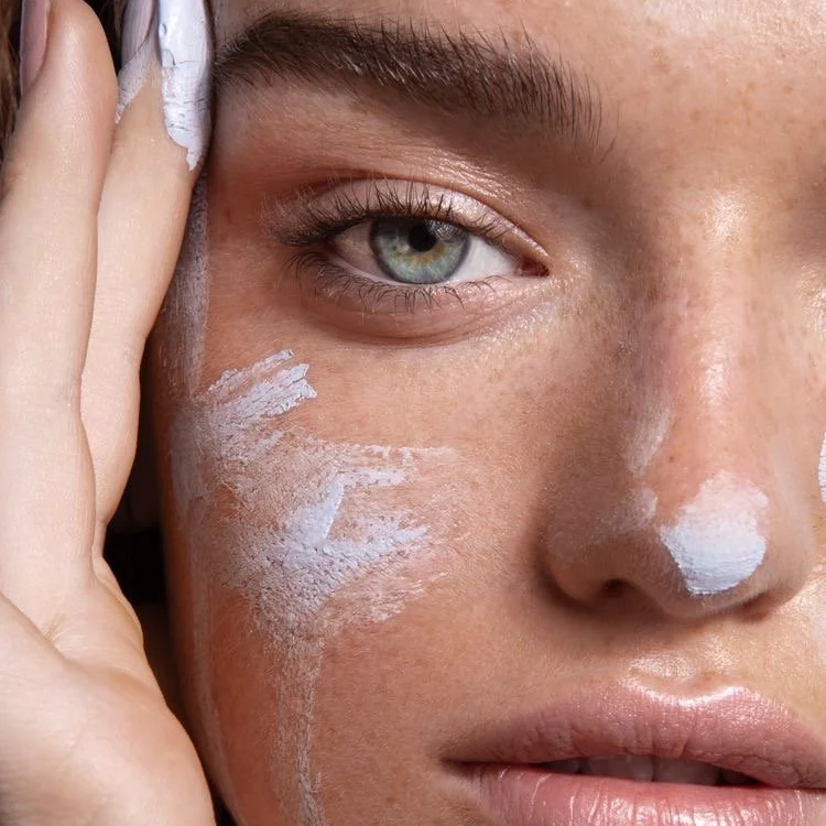 A close-up of a woman's face with white cream applied to her cheek and nose, touching her temple with her hand.