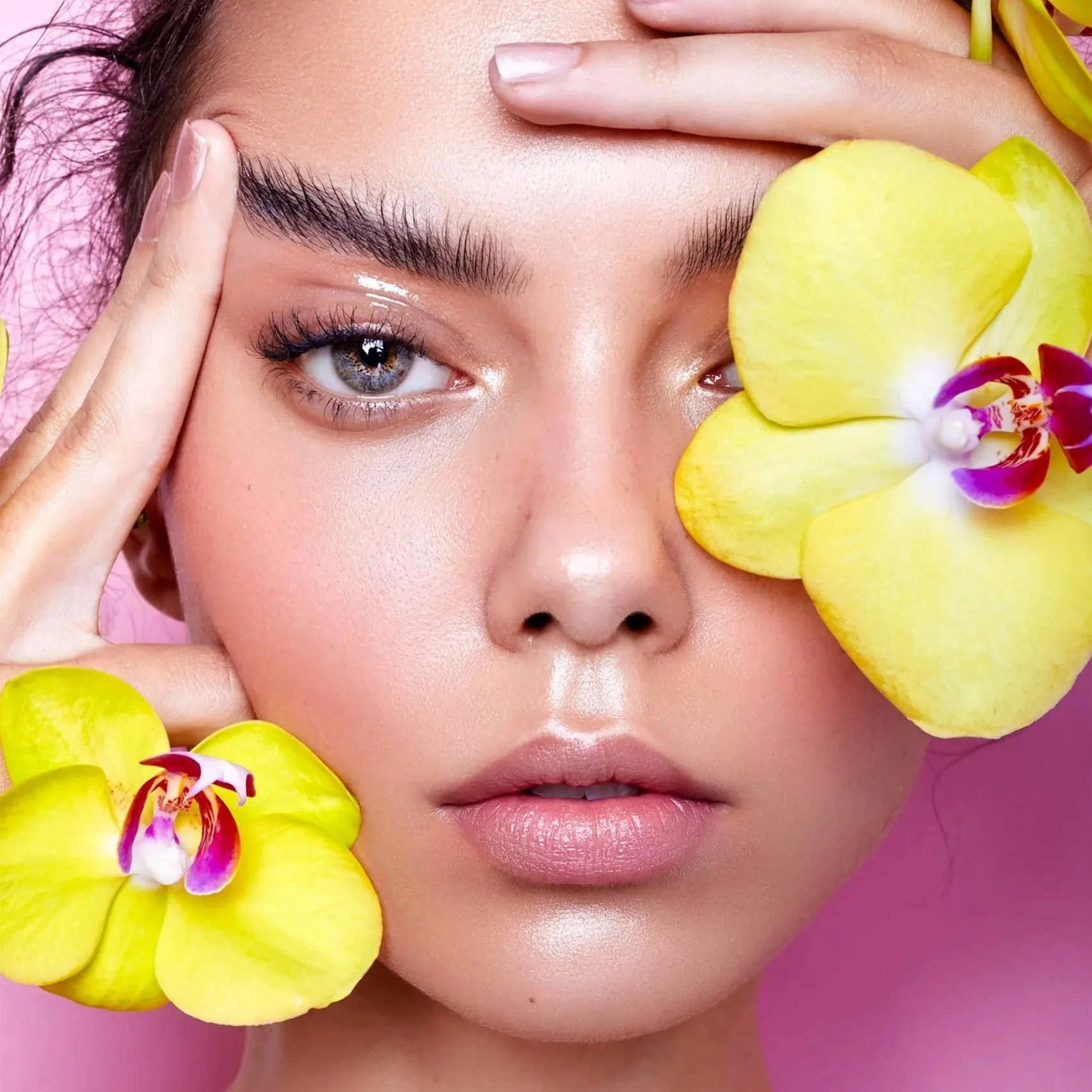 Close-up of a woman's face surrounded by yellow orchid flowers with pink and purple accents, with her hand gently touching her forehead and her eye, looking directly at the camera against a pink background.