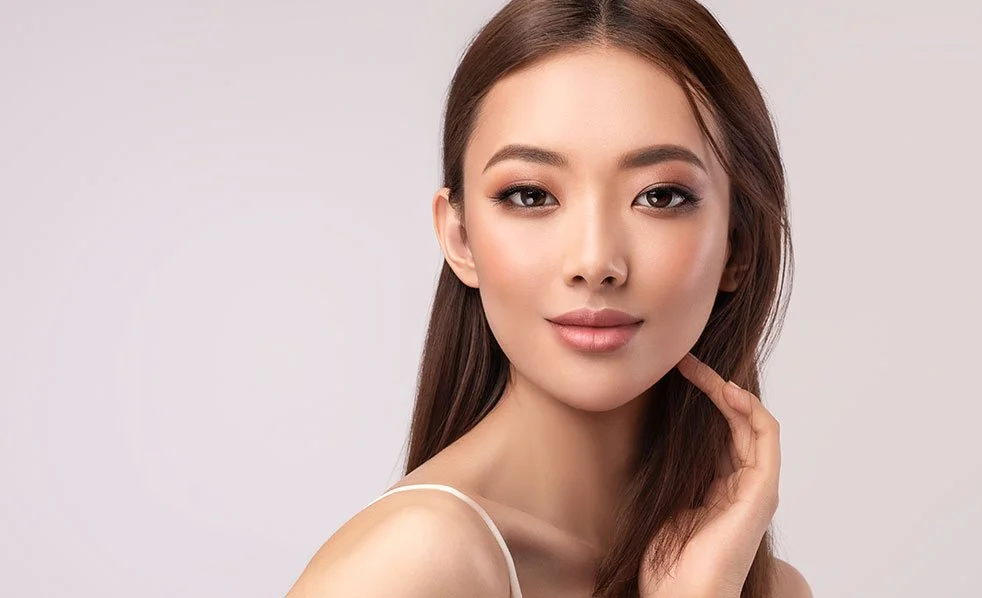 A portrait of a young woman with long brown hair, wearing subtle makeup, and a light-colored top, posing in front of a plain background.