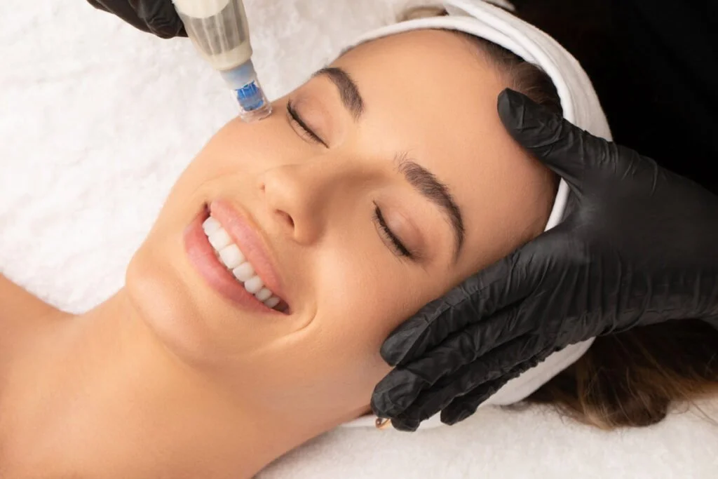 A woman receiving a cosmetic facial treatment with a syringe near her eye, lying down with her eyes closed and smiling, while a practitioner's hand in black glove supports her head.