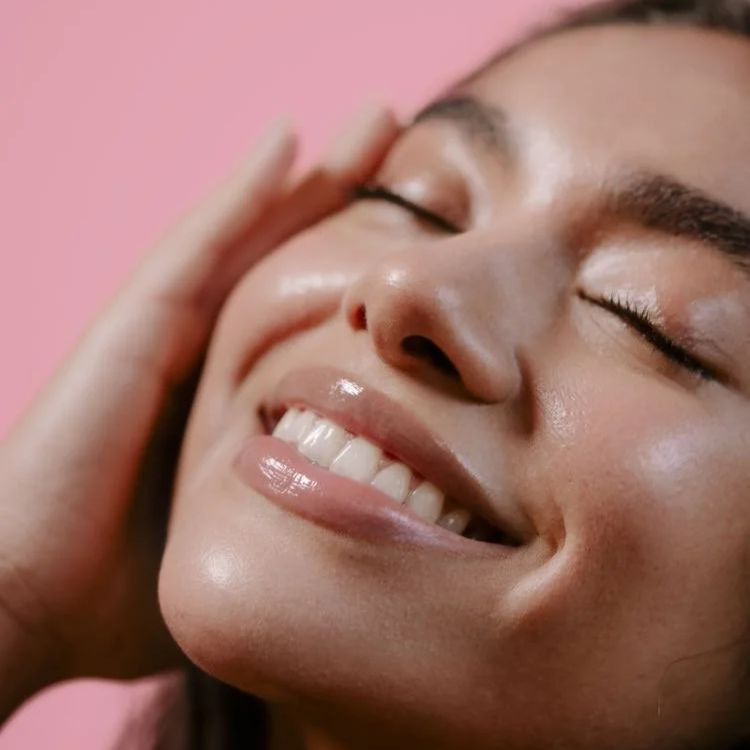 Close-up of a woman smiling with her eyes closed, touching her face, against a pink background.