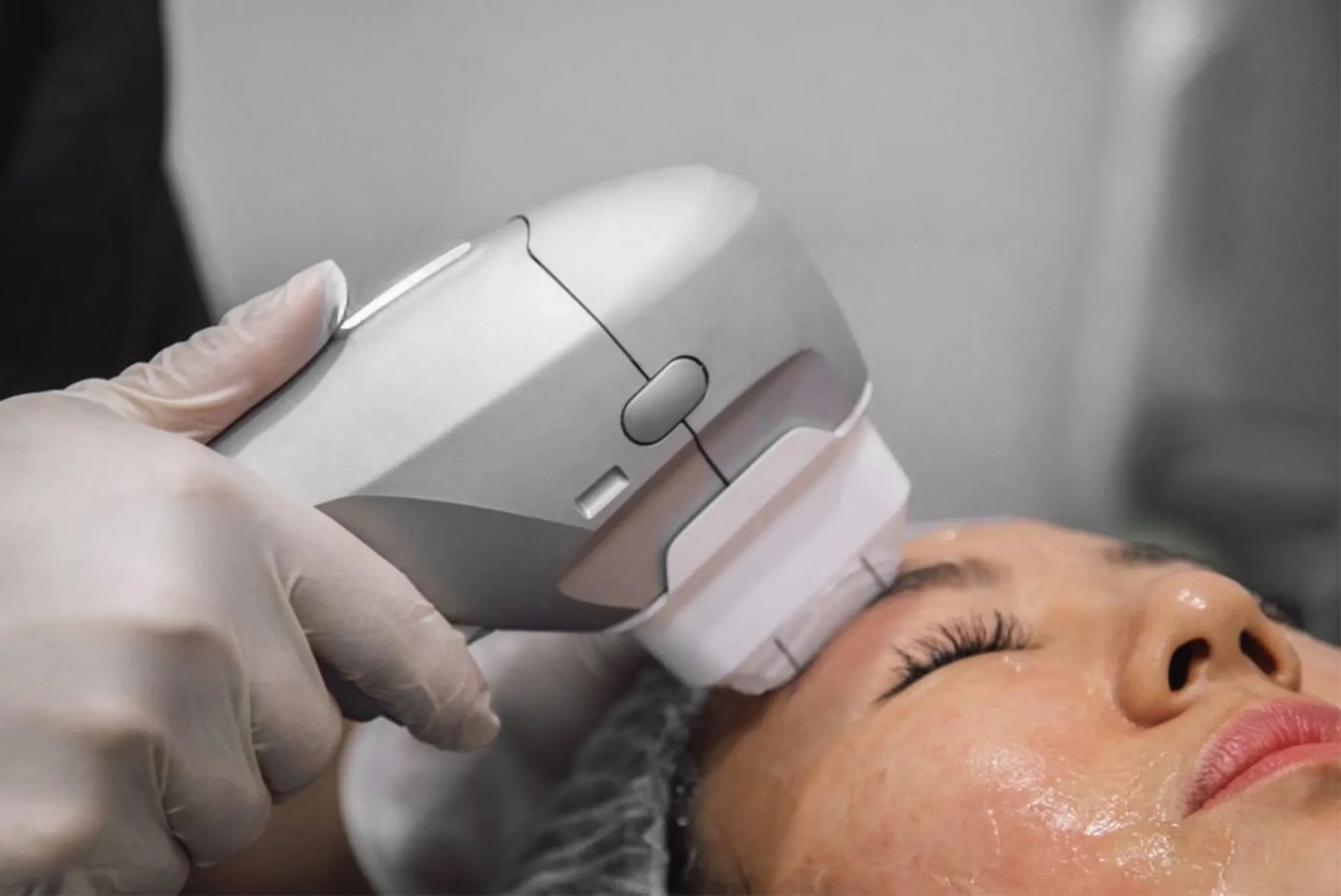A person with closed eyes receiving a cosmetic or dermatological treatment from a professional wearing gloves, using a device near their face.