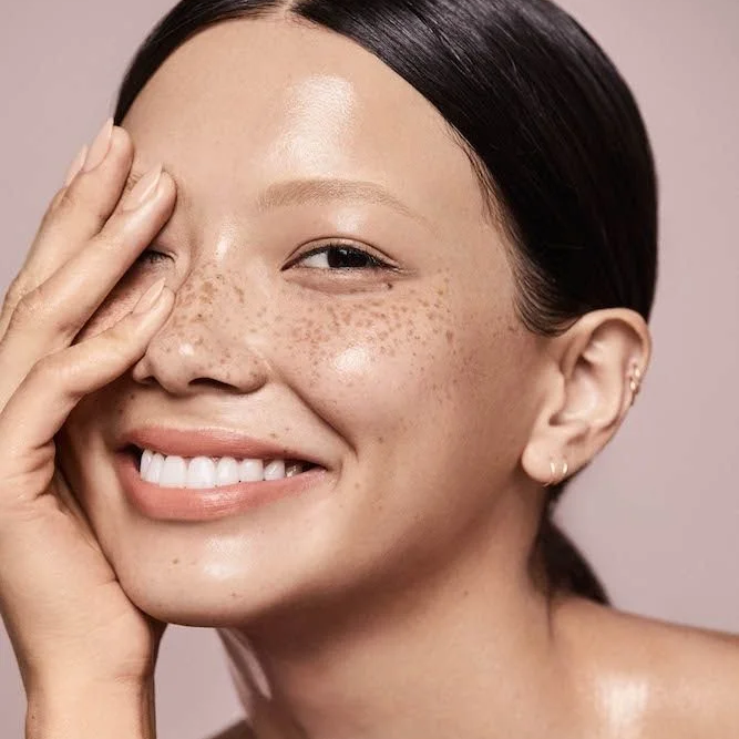 Close-up of a smiling woman with freckles, holding her face with one hand, against a soft pink background.