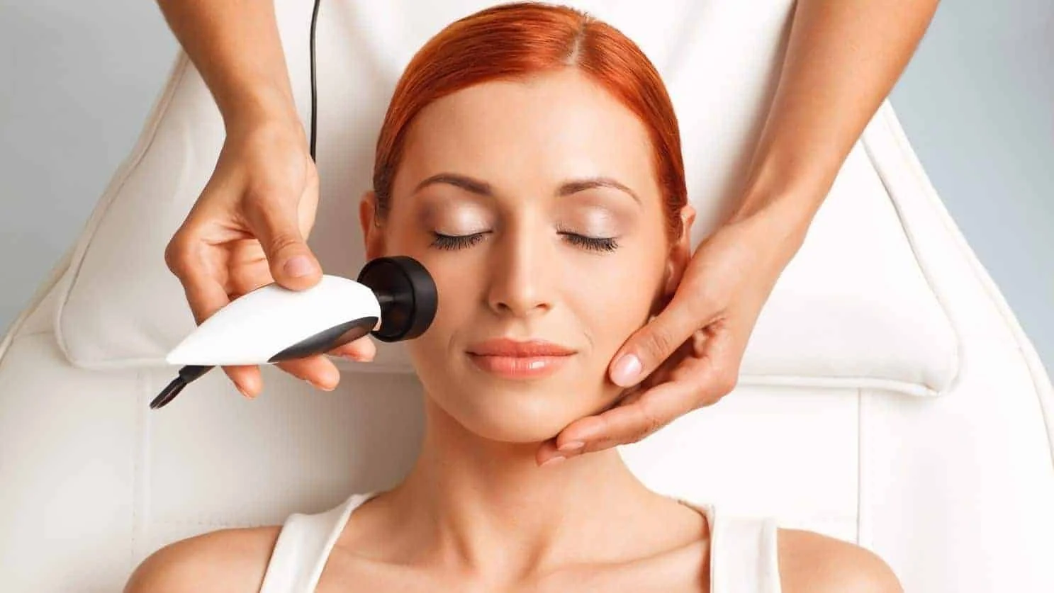 A woman receiving a facial treatment with a handheld device while lying on a treatment bed, with her eyes closed and a relaxed expression.