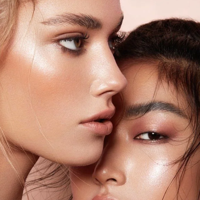 Close-up of two women with luminous skin, one with blonde hair and the other with dark hair, positioned very close together, displaying makeup with a focus on their eyes, eyebrows, and lips.