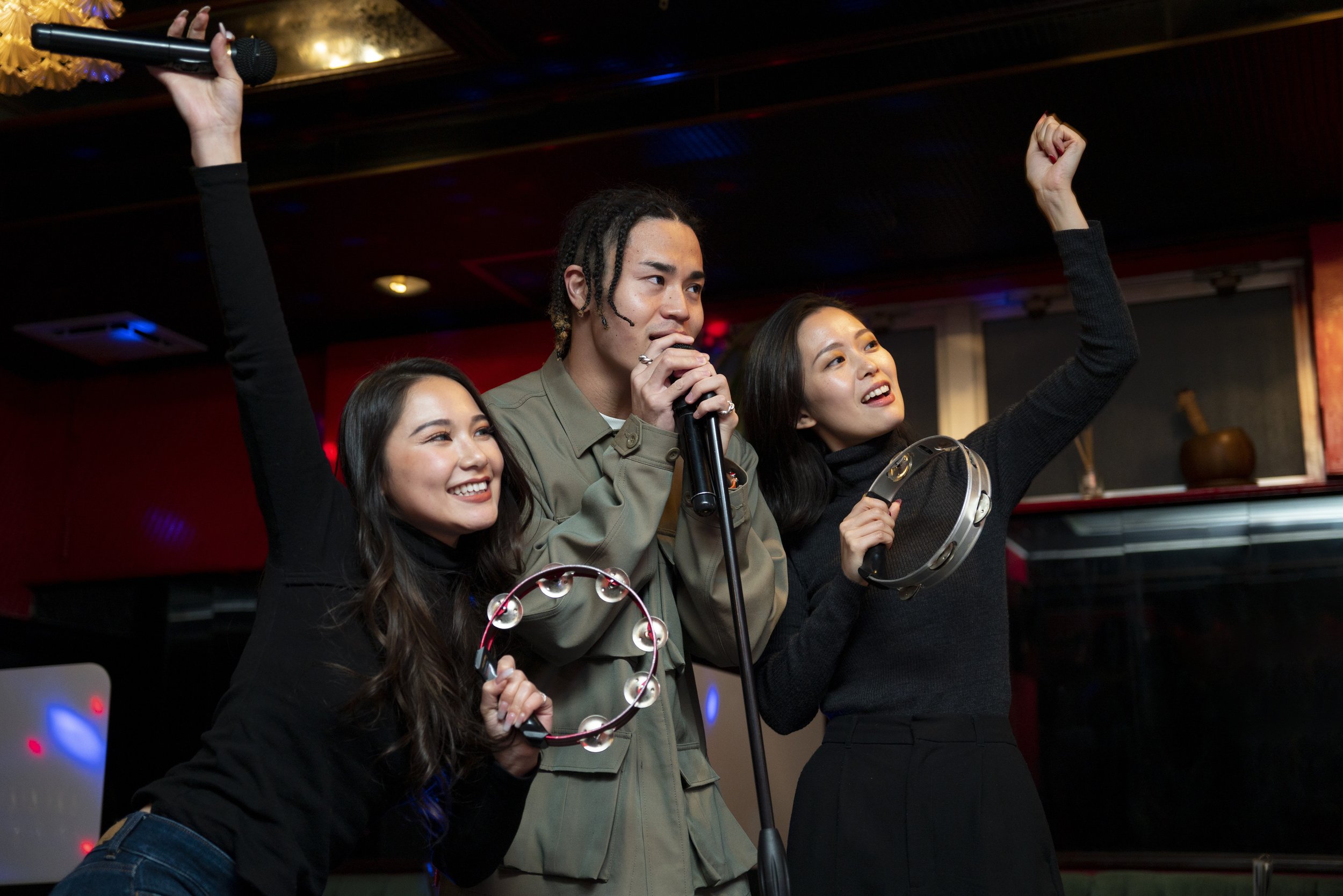 Three people singing and dancing at a karaoke party with musical instruments, one holding a microphone, in a dimly lit room with colorful lighting.