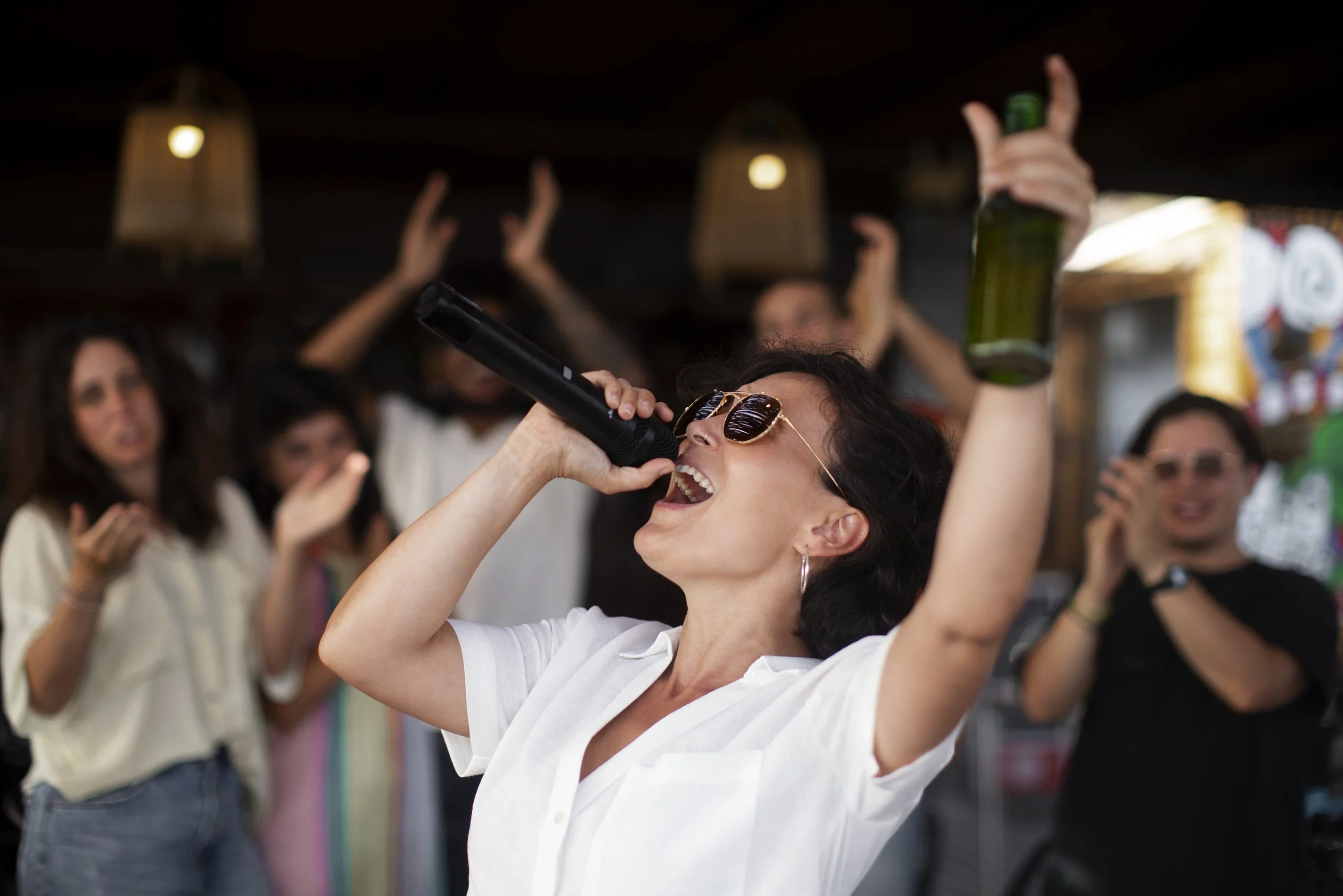 A woman singing into a microphone at a lively social gathering, holding a green bottle in her other hand, with a crowd of people around her.