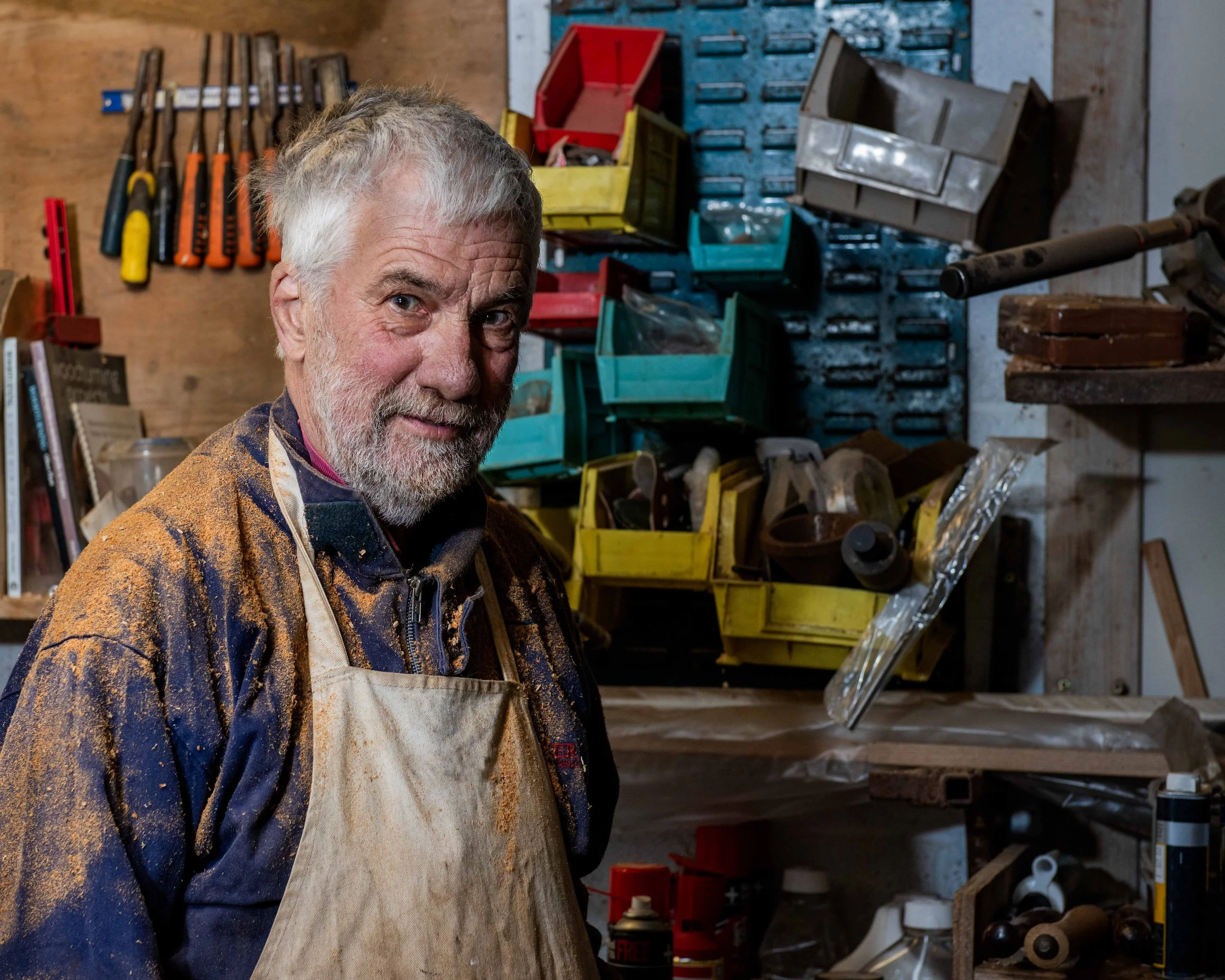 An elderly man with gray hair and a beard, wearing a colorful jacket and a dirty apron, standing in a woodworking workshop with tools, plastic storage bins, and woodworking materials in the background.