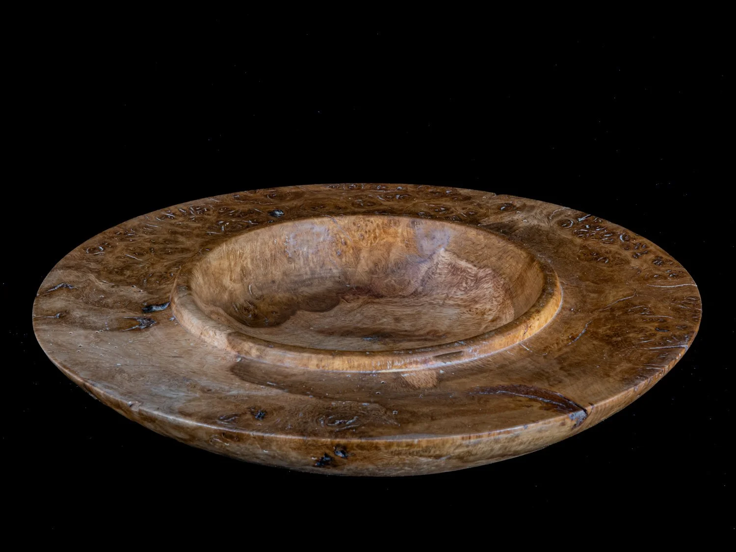 A round wooden bowl with a shallow, carved interior surface placed against a black background.