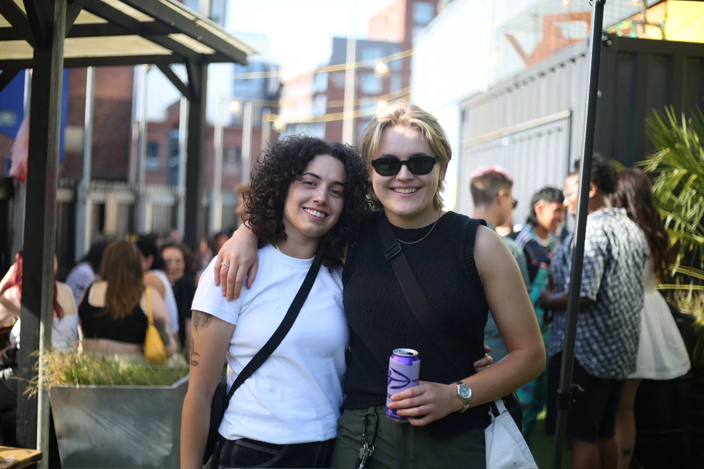 Two women smiling and embracing each other at an outdoor gathering or festival, with people and string lights in the background.