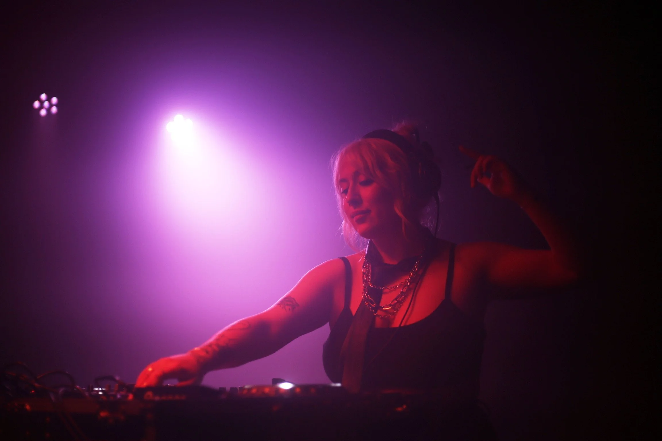 A female DJ with blonde hair and tattoos, wearing headphones and a black outfit adorned with chains, performing at a nightclub with pink and purple lighting.