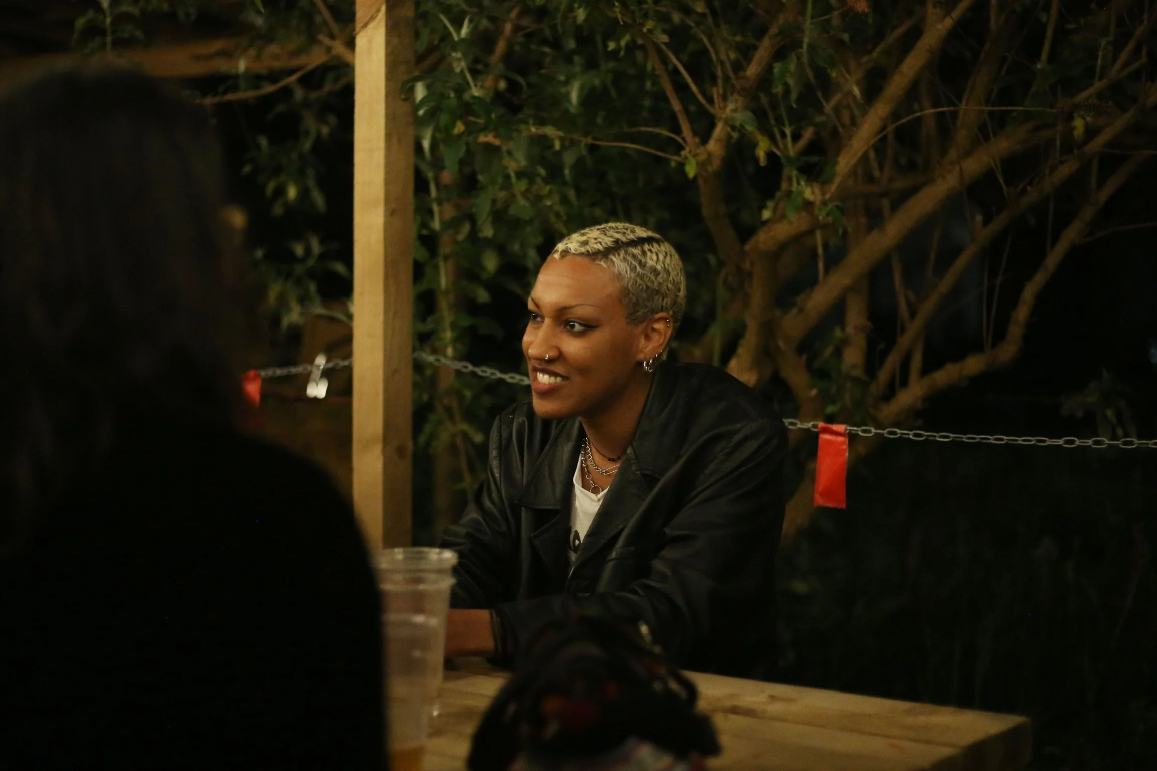 A person with short, platinum blonde hair and multiple earrings sitting outdoors at night, smiling and talking to others, with trees in the background.