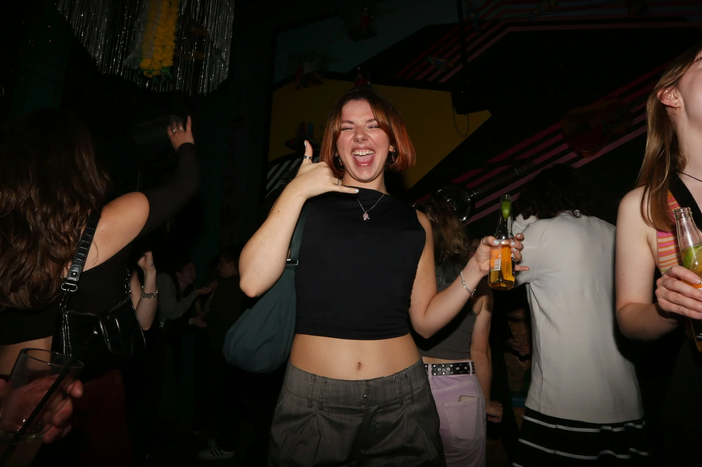 A young woman with red hair smiling and making a phone gesture with her hand at a party or club. She is holding a beer bottle and is wearing a black crop top and a necklace. Other people are around her, some holding drinks, in a dark, festive environ