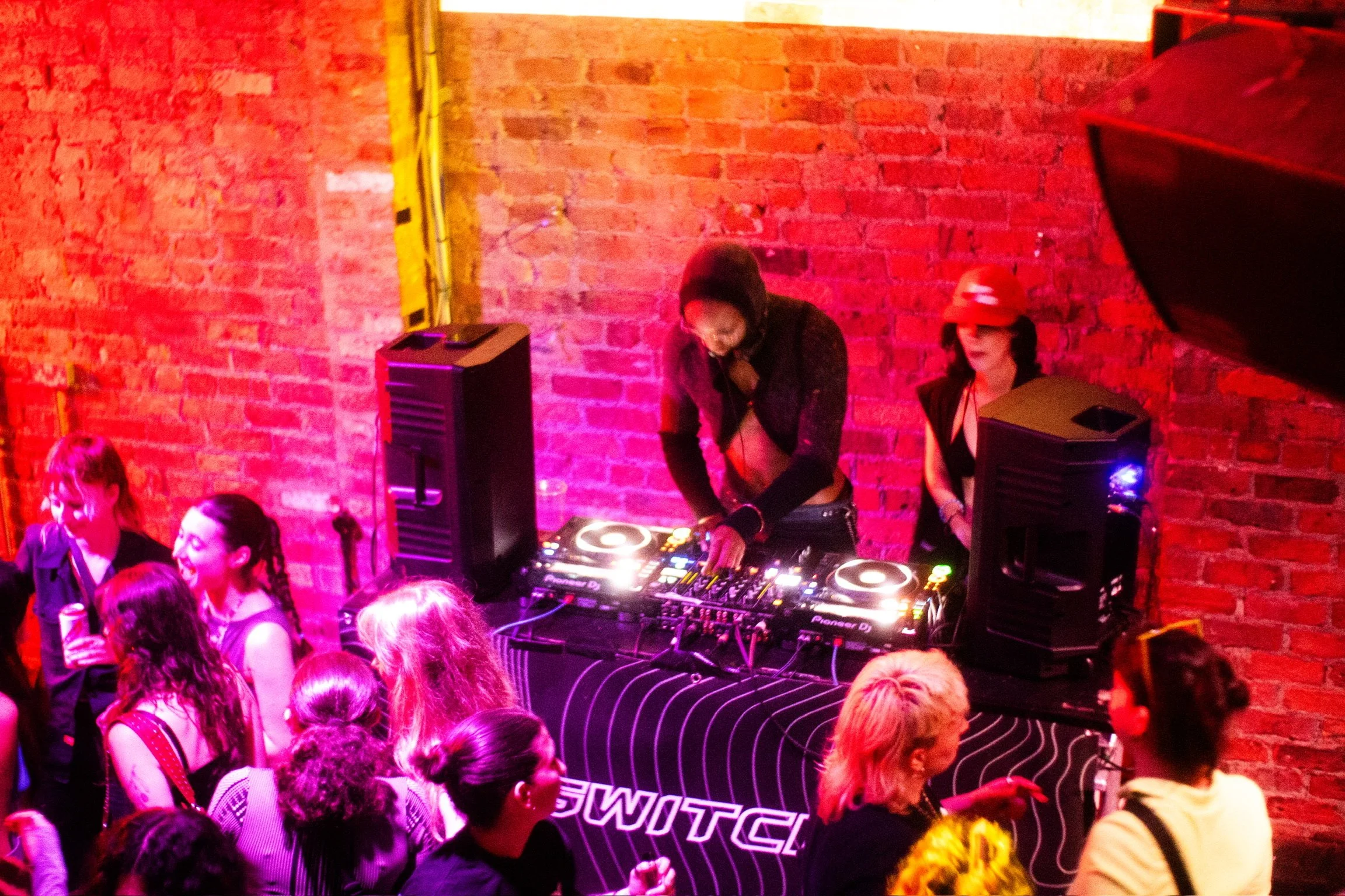 A DJ is performing at a party with a crowd of people dancing in front of him. The DJ is using a Pioneer DJ controller and is wearing a hooded jacket. The background features a red brick wall, illuminated with pink and purple lighting.