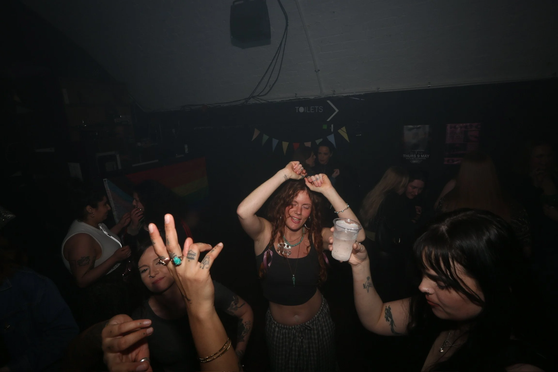 People dancing at a party with a woman holding a drink in the foreground. The background has a sign pointing to toilets and colorful bunting decorations, with a dark, lively atmosphere.
