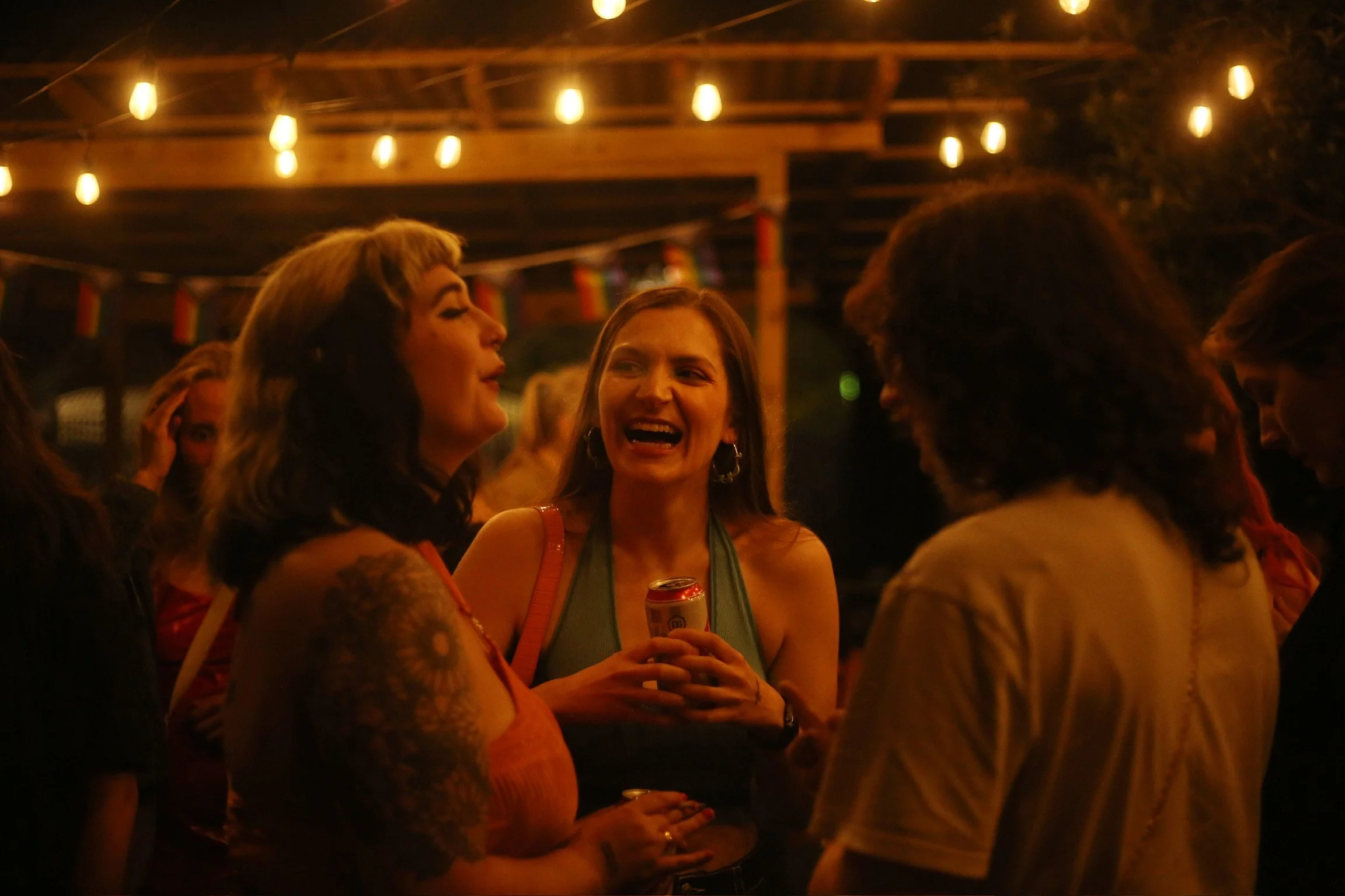 Group of women having conversations at an outdoor party during the evening, under string lights, with one woman holding a can of beer.