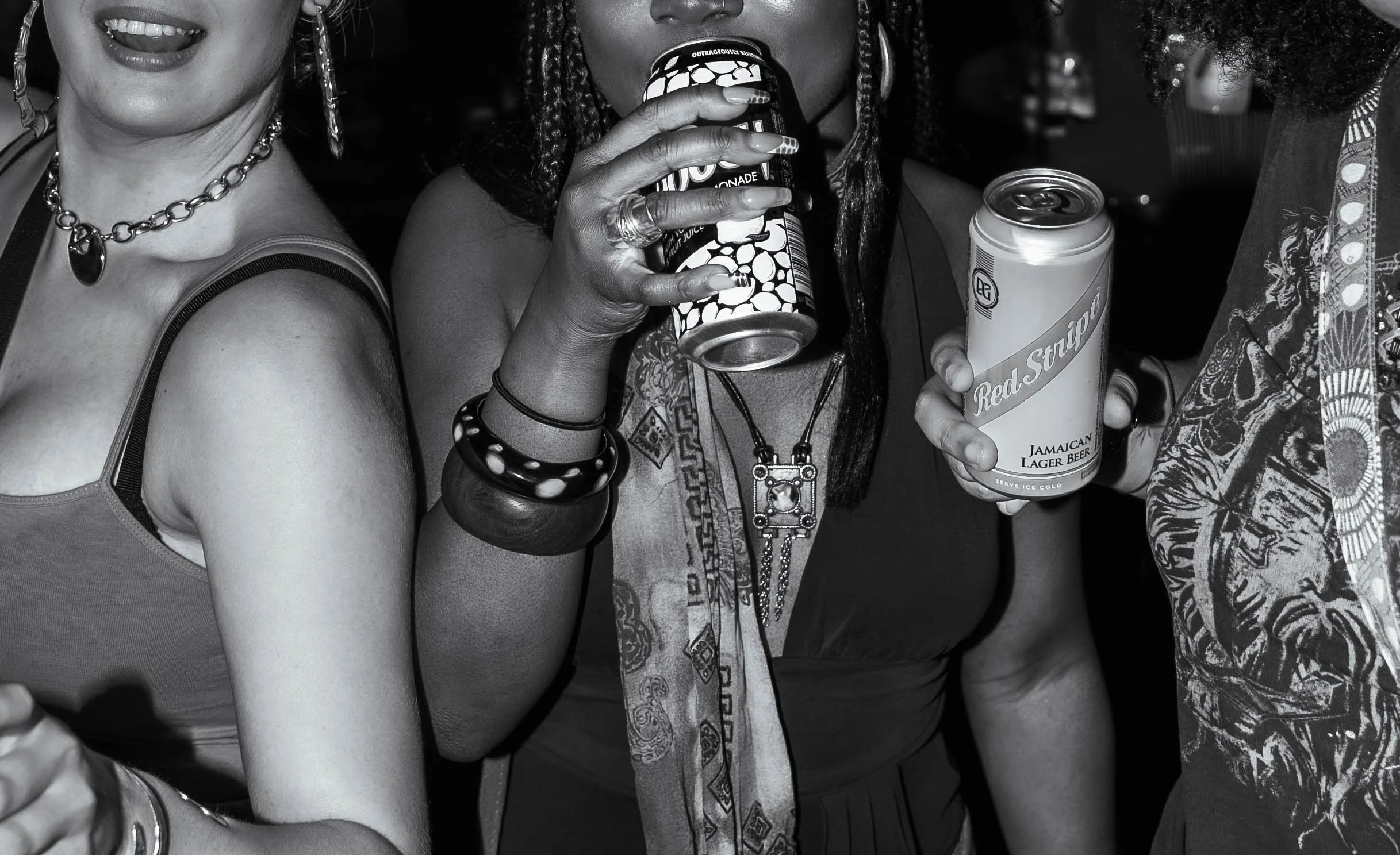 Close-up of three women holding canned drinks, with only their torsos visible. The woman in the middle is sipping from a can with a pattern, holding it with her right hand, and wearing multiple necklaces and bracelets. The woman on the left is wearin