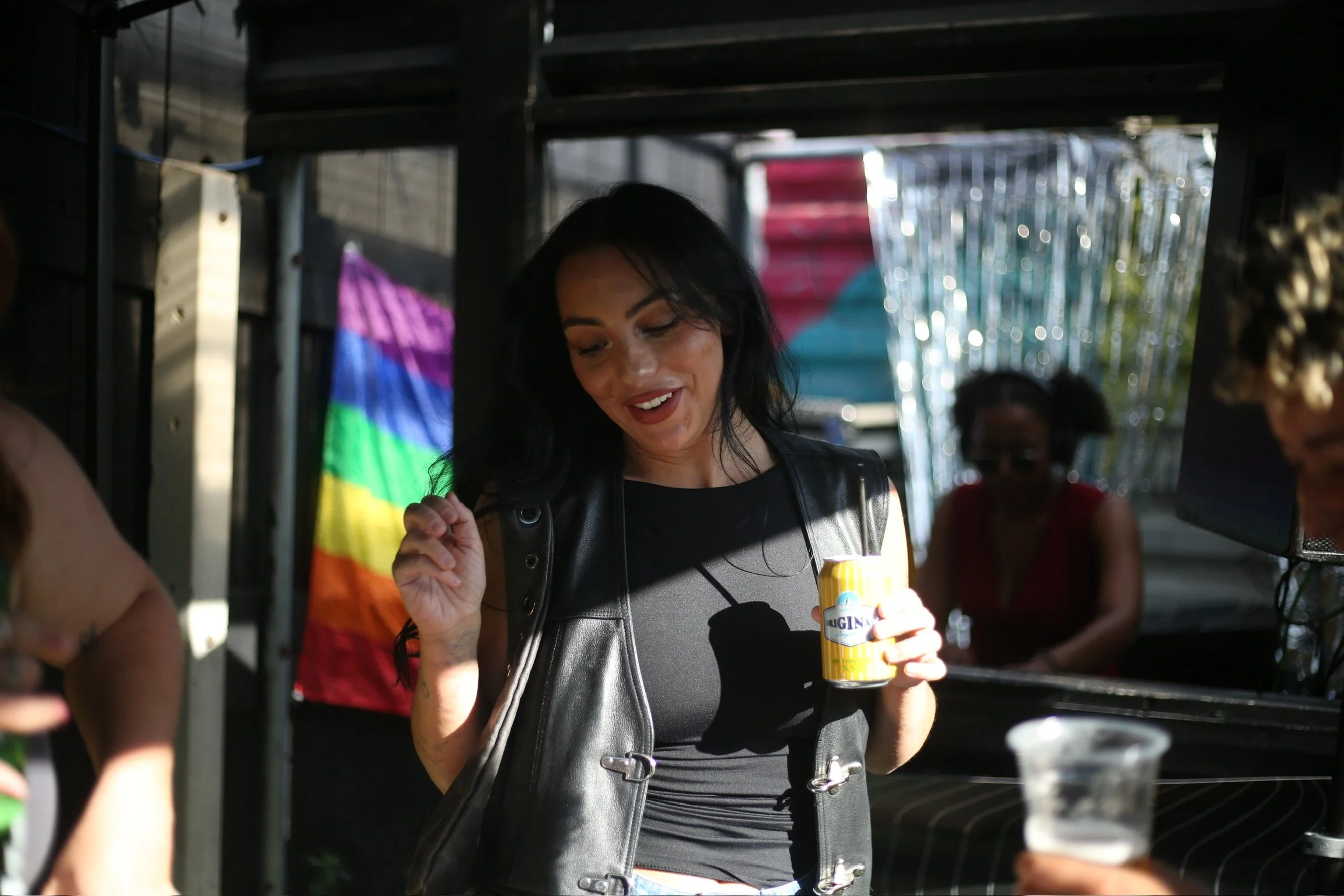 A woman with black hair and a black leather vest holding a yellow and white container, standing among other people under a dark structure with rainbow pride flags and shiny decorations in the background.