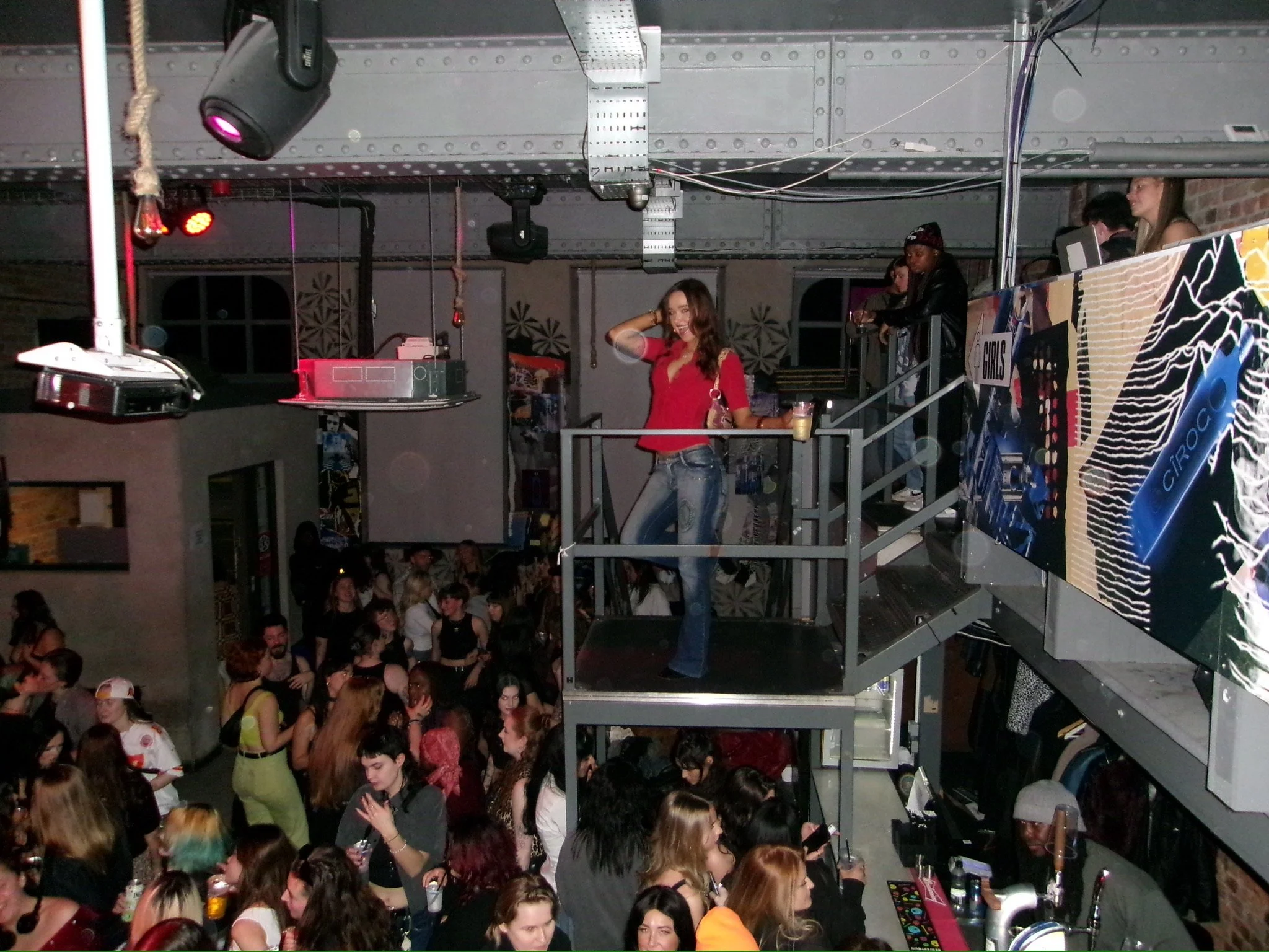 A crowded indoor party or nightclub with people dancing and socializing. There is a woman in a red shirt smiling and posing in the middle, holding a cup, with a drink. The background shows a DJ booth with equipment, people on an upper level, colorful