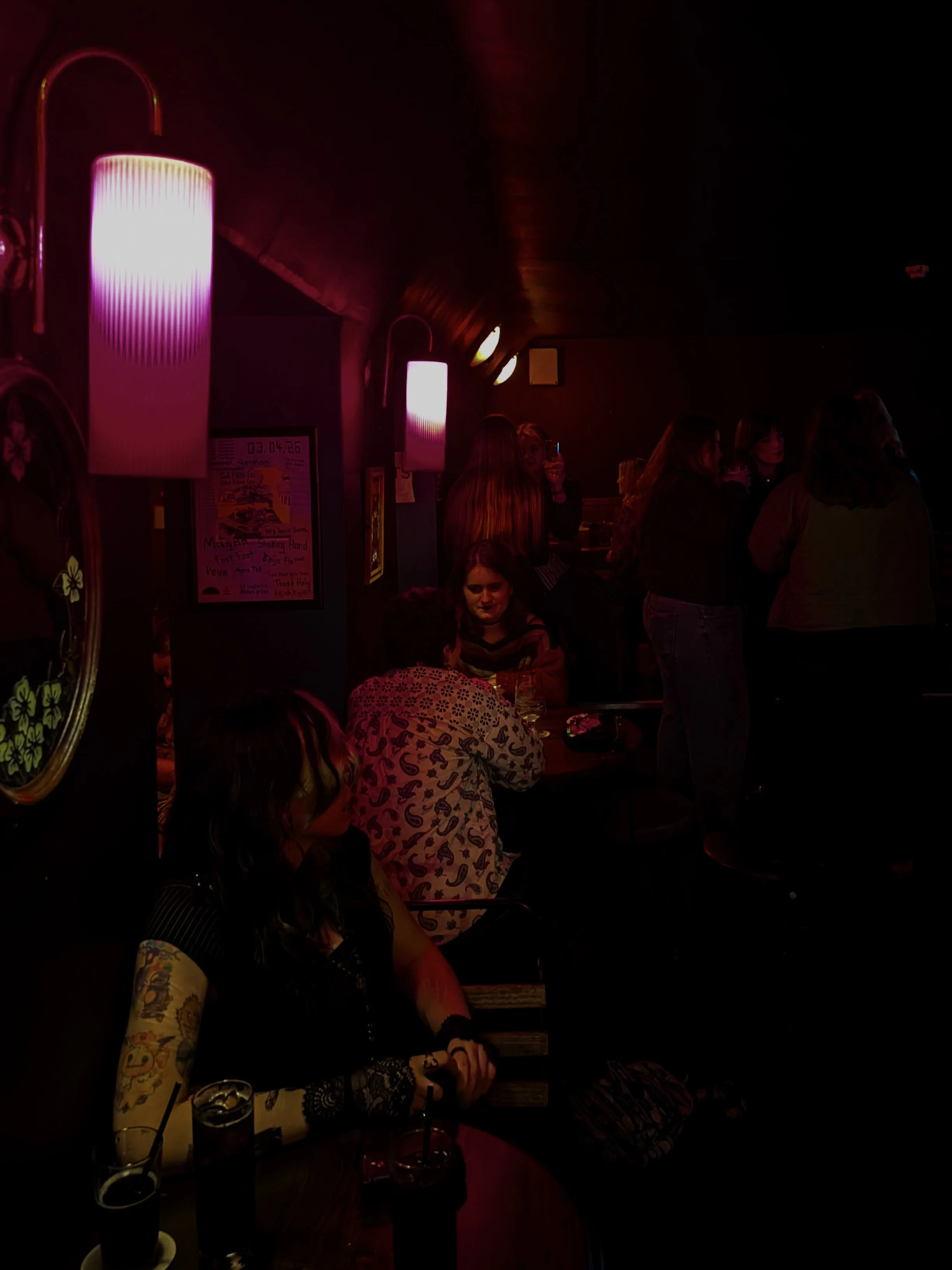 A dimly lit bar or restaurant with pink neon or mood lighting, several people socializing and standing around, with drinks on the tables. There are wall-mounted lights and various posters on the wall.